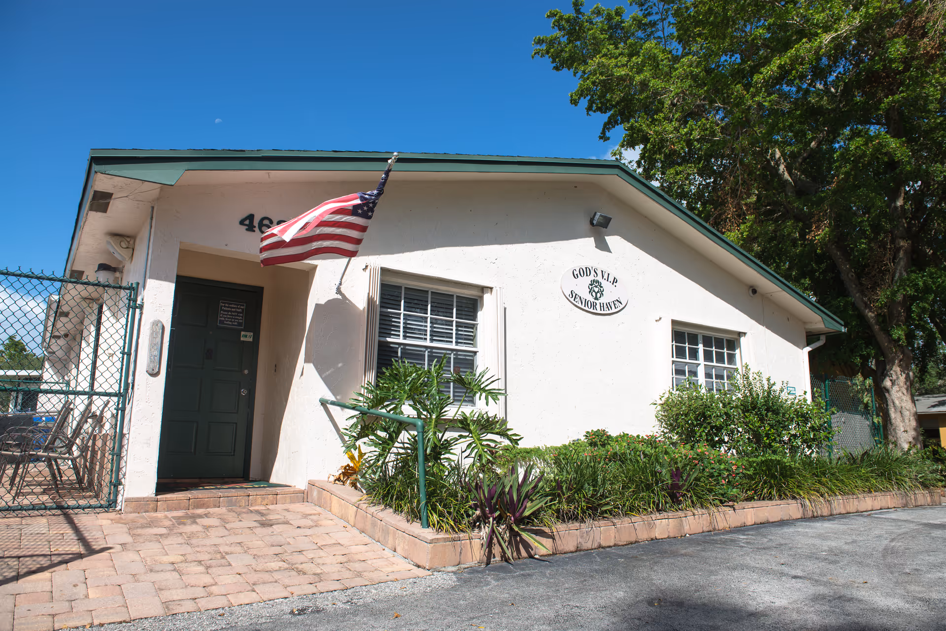Exterior view of a single-story building with a green roof and white walls. An American flag is mounted near the entrance door, which is dark green. There are two windows with white shutters and a garden with various plants in front of the building. A sign on the wall reads 'GOD'S V.I.P SENIOR HAVEN'. The sky is clear and blue, and there are trees in the background.