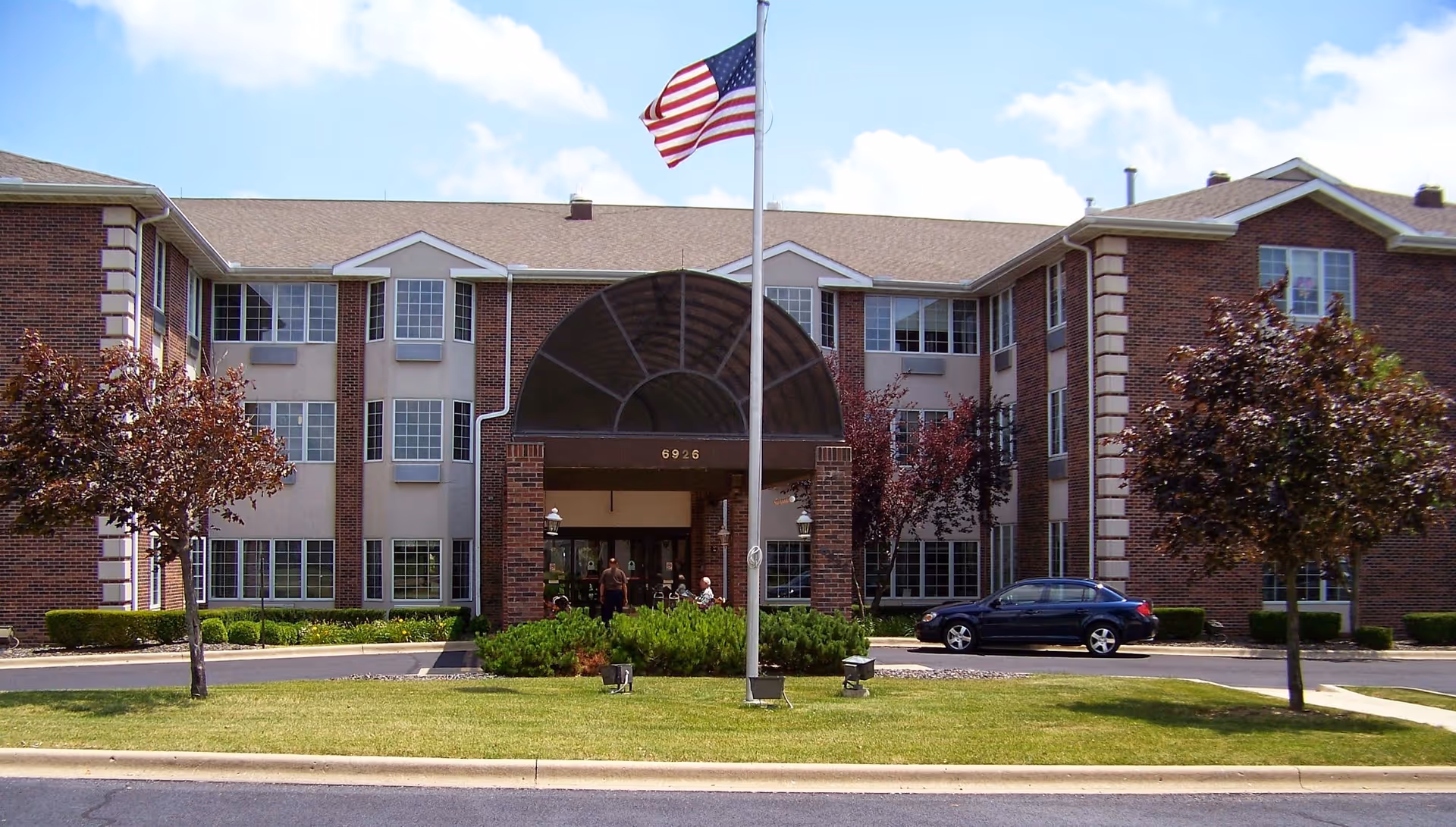 Exterior view of Manor Court of Peoria senior living facility with flag, entrance, and landscaping.