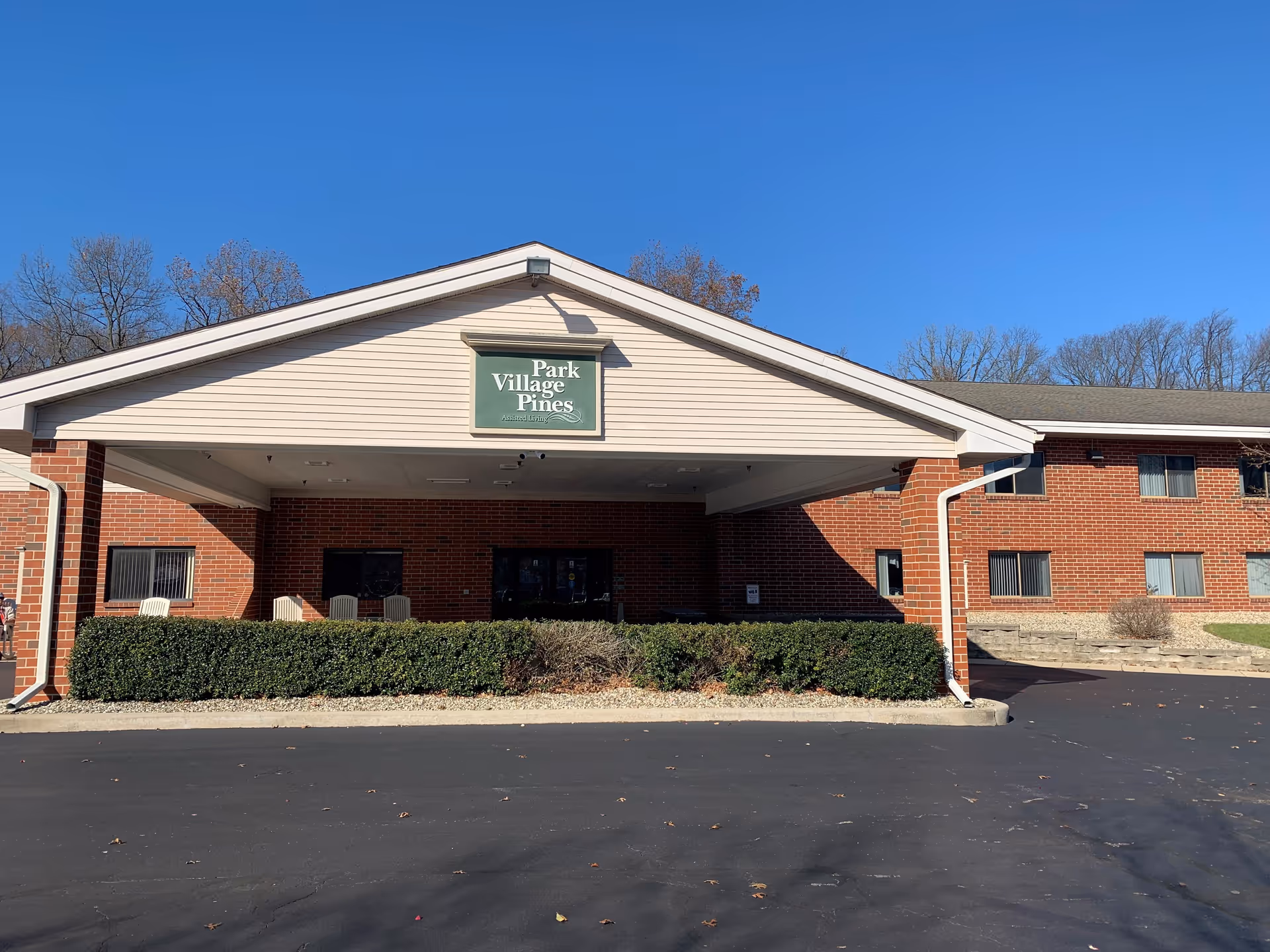 Exterior front view of Park Village Pines assisted living facility with a covered entrance, brick walls, and a clear blue sky.