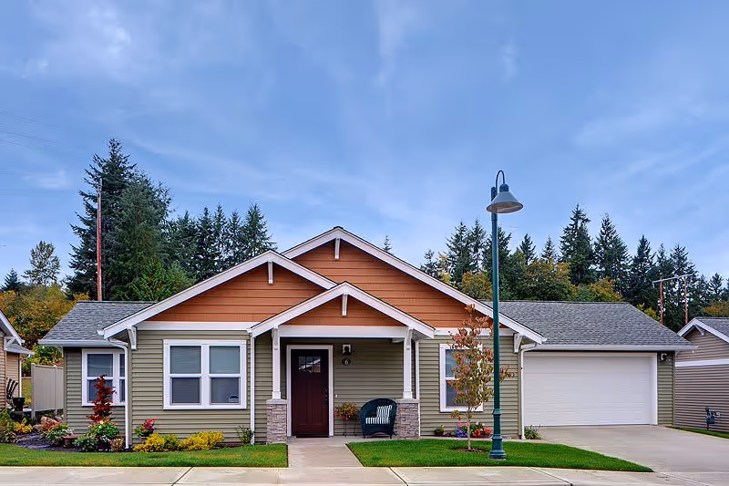 Single-story ranch-style house with a front porch, attached garage, lamp post, and landscaped lawn under a cloudy sky.