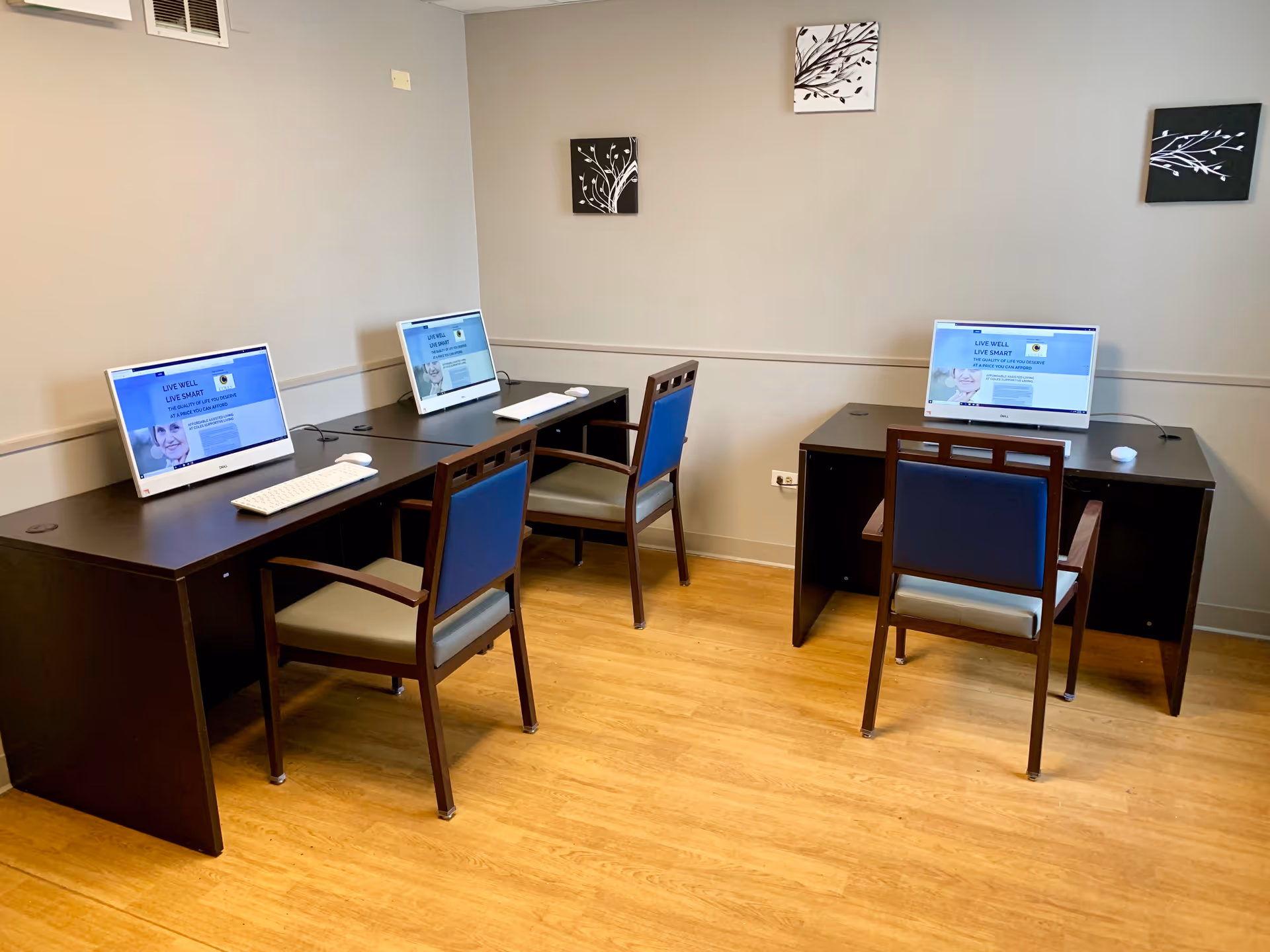 Computer workstation area with three desks, monitors, and blue chairs inside a senior living facility.