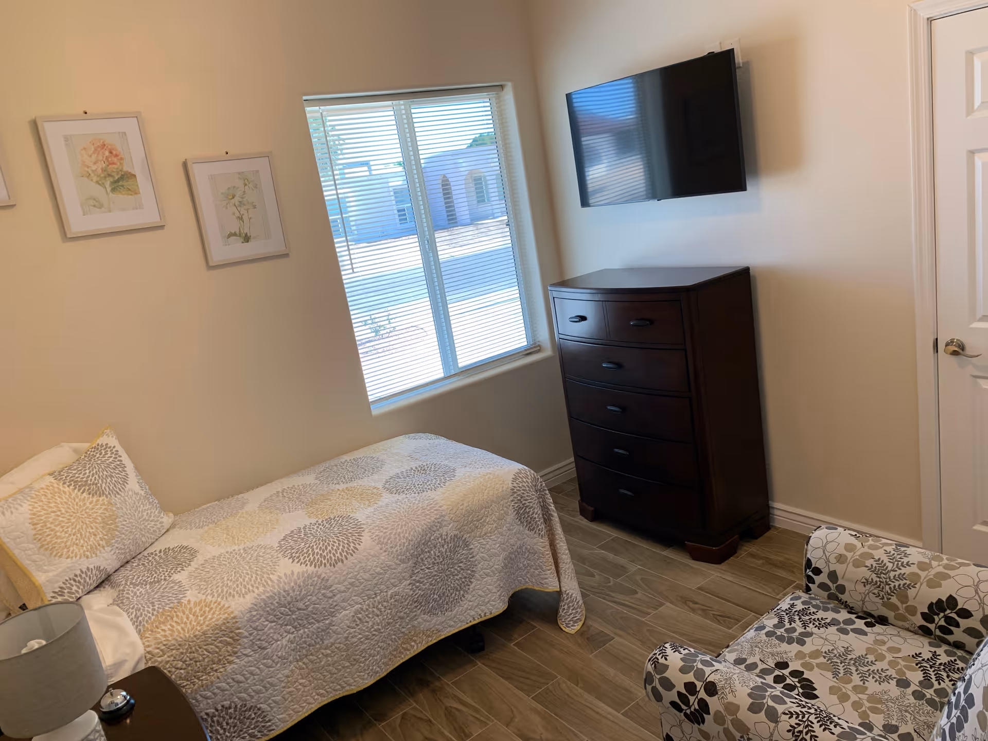 A small bedroom with a single bed covered in a patterned quilt, a bedside table with a lamp, a floral-patterned armchair, a dark wooden chest of drawers, a wall-mounted flat screen TV, and three framed botanical prints on the wall. A window with blinds lets in natural light.