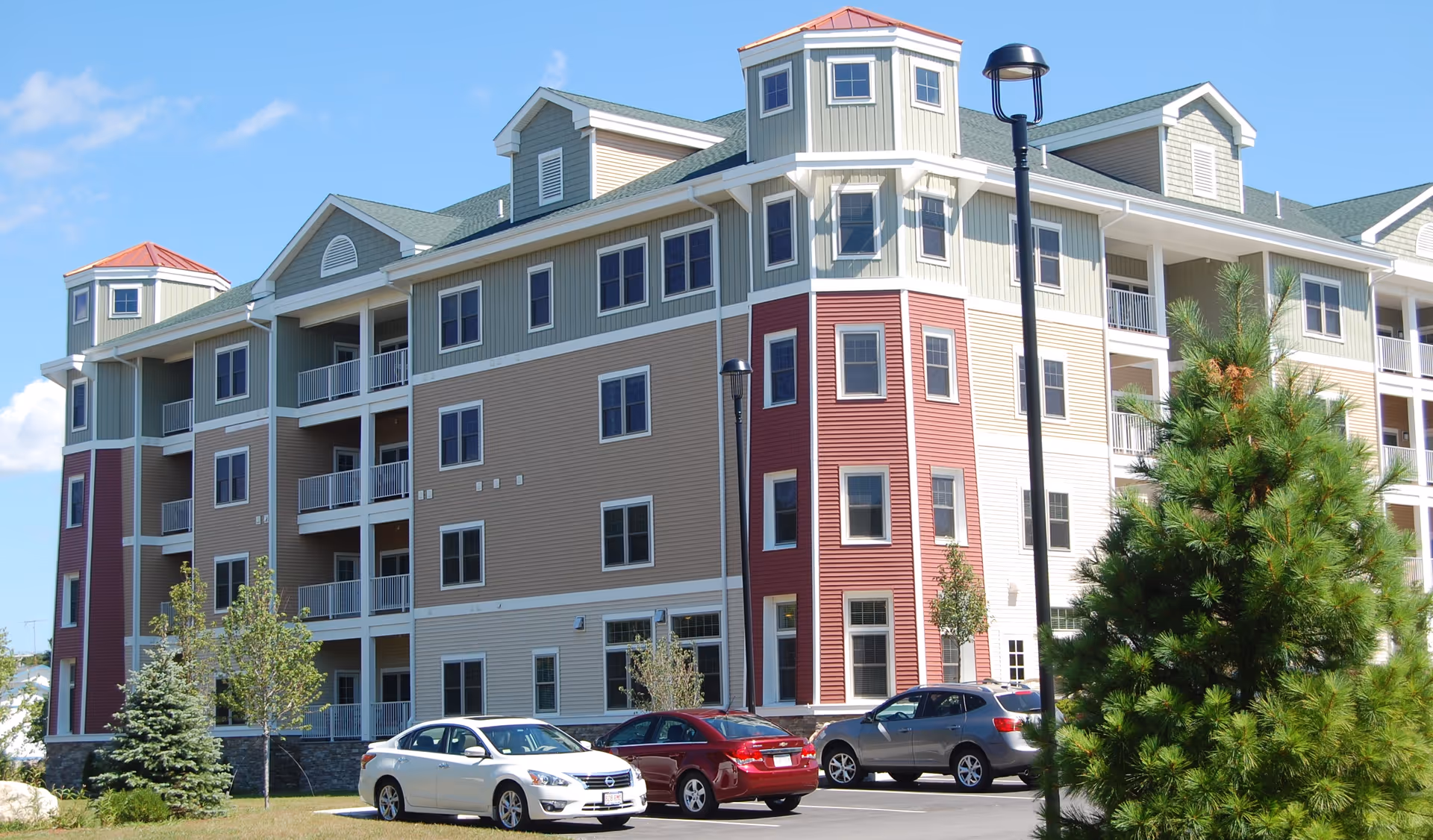 Exterior view of a multi-story senior living facility building with beige, red, and light green siding, multiple windows, balconies, and a parking lot with several cars in front. There are trees and a clear blue sky in the background.