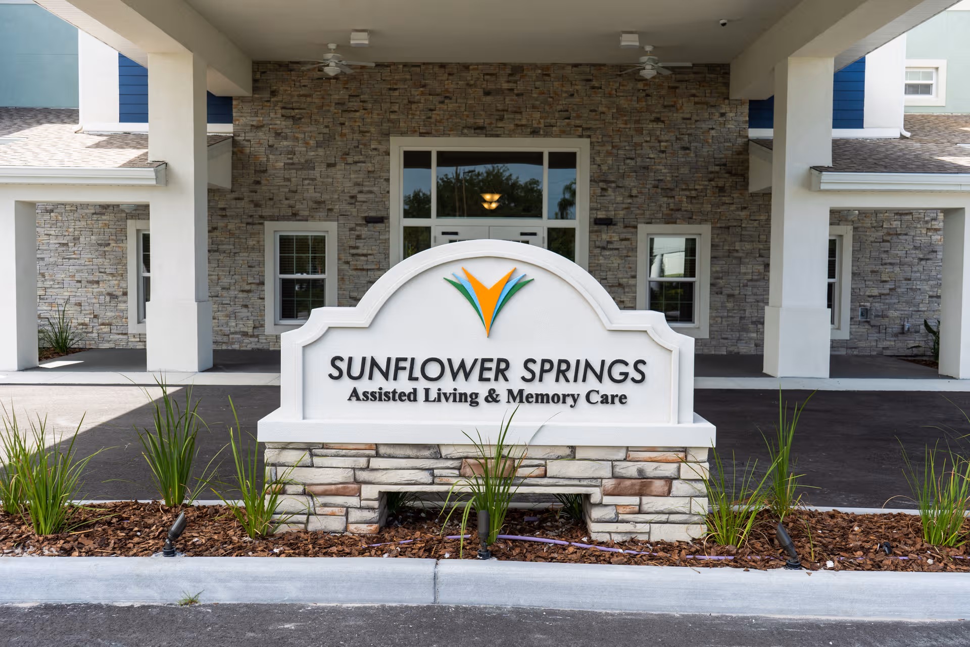 Front exterior of Sunflower Springs Assisted Living at Trinity with a stone sign and landscaping.