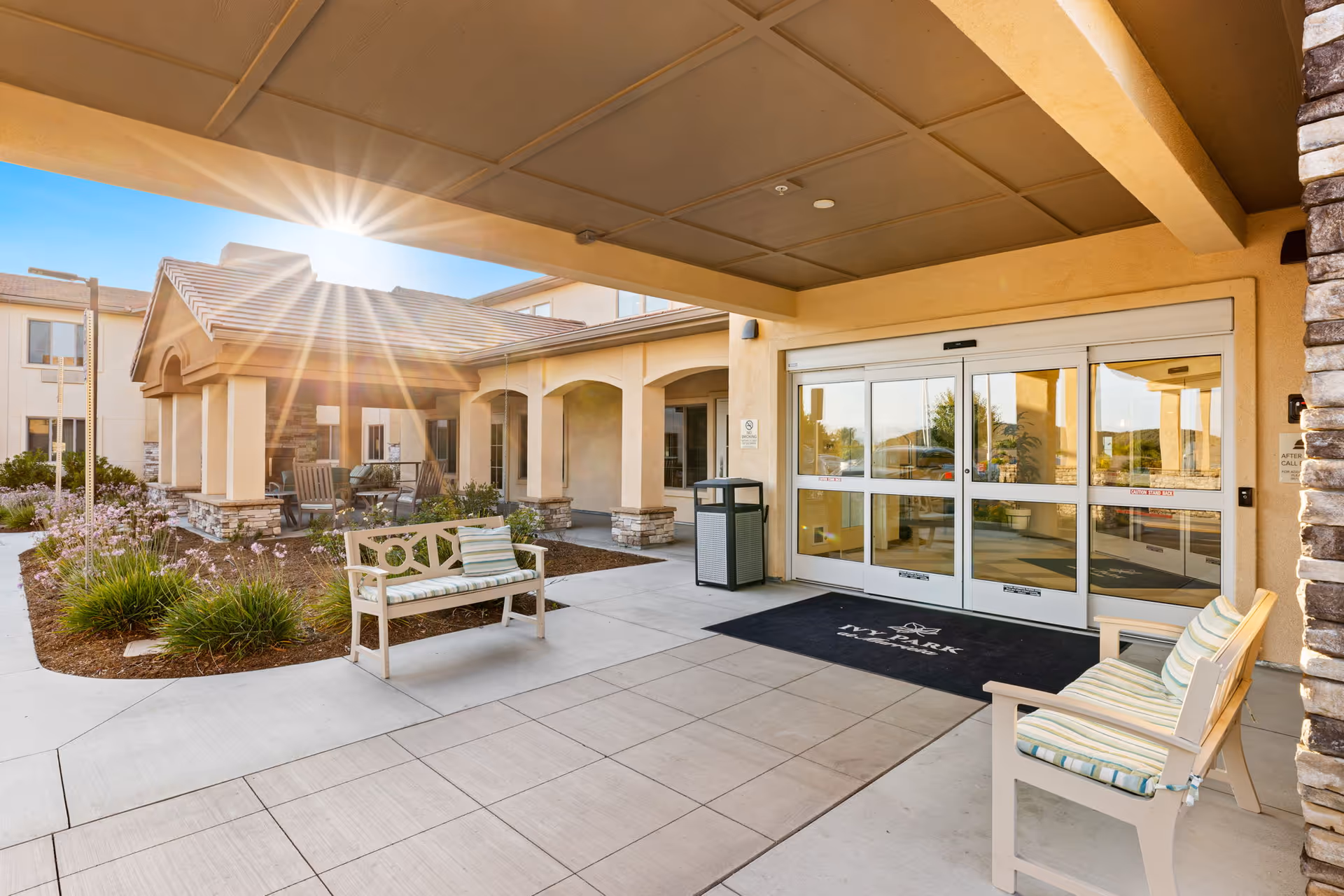 Covered entrance to a senior living facility with benches, landscaping, and automatic sliding glass doors under a sunny sky.