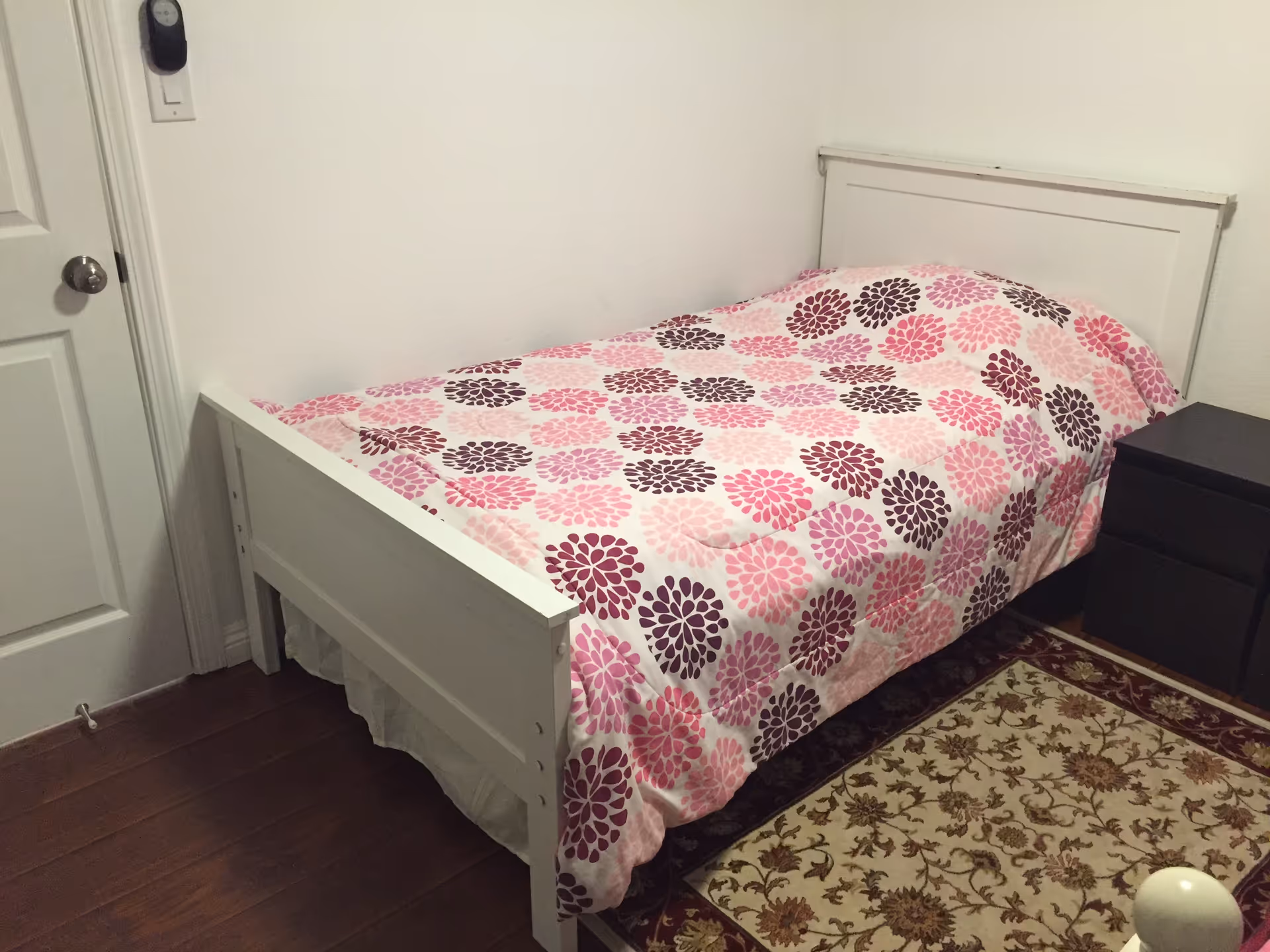Small bedroom with white bed frame, pink floral quilt, nightstand, and patterned rug.