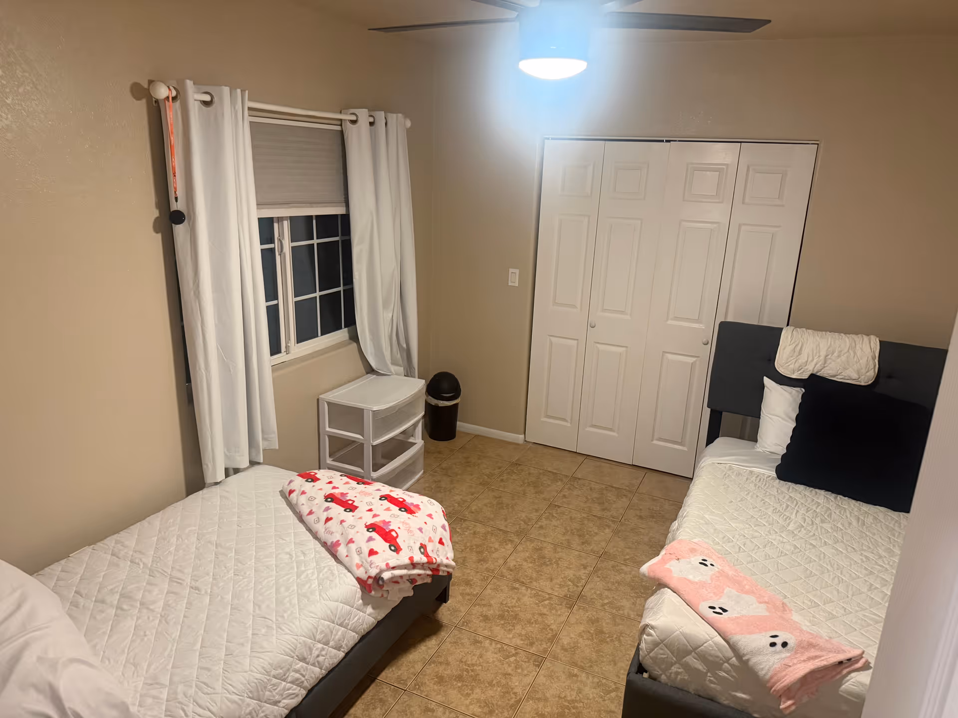 Two single beds with white quilts sit in a beige bedroom with a window and closet.