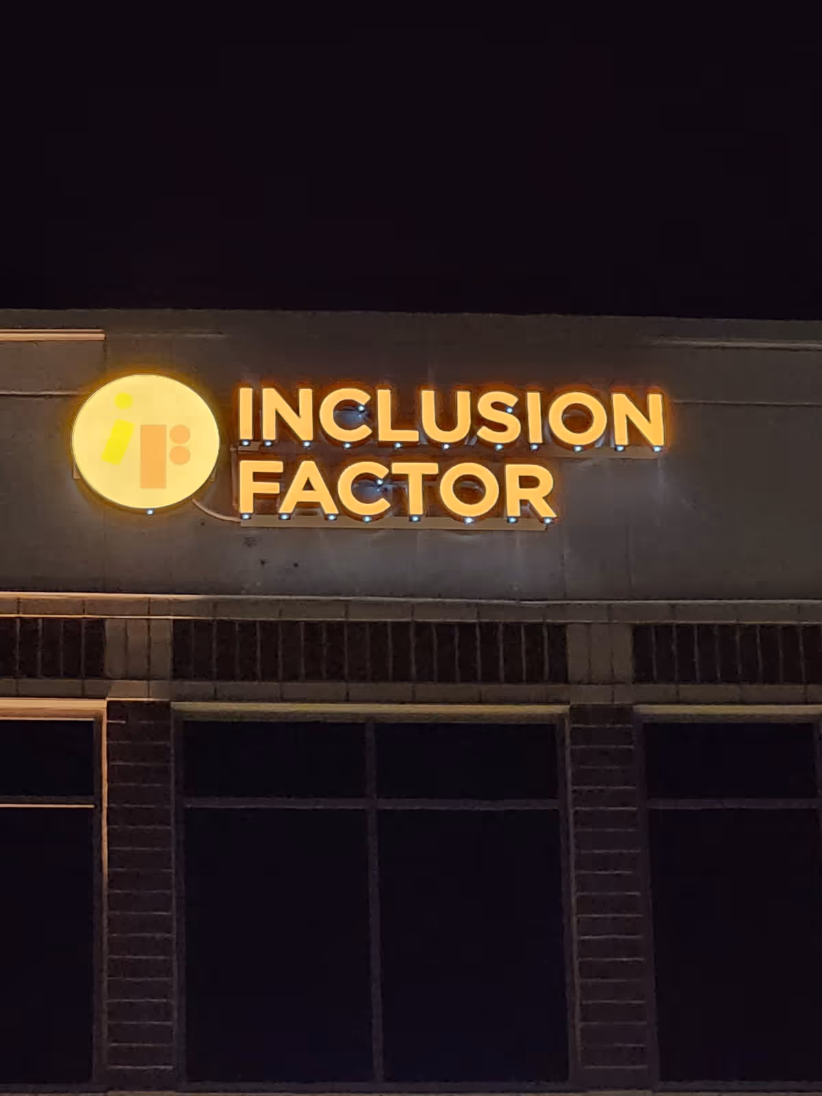 Nighttime exterior of a building with a glowing INCLUSION FACTOR sign and circular logo.