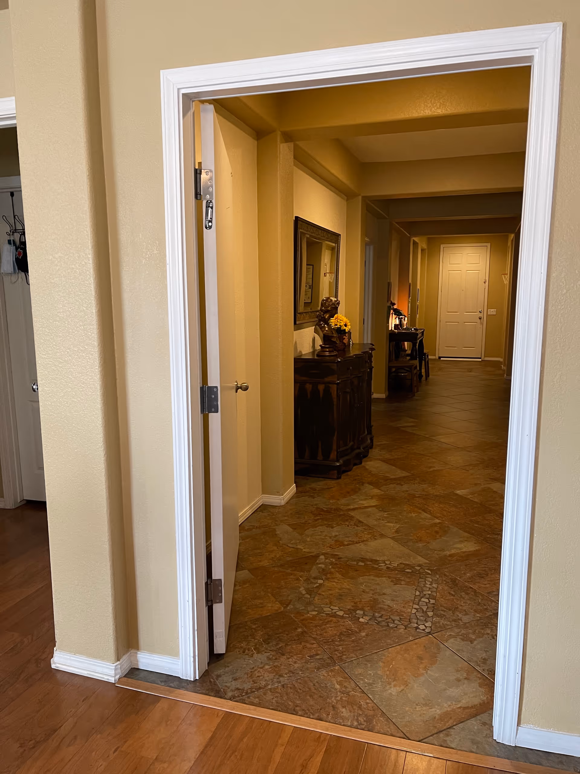 View through a doorway into a hallway with tiled floor and beige walls. The hallway features a decorative wooden cabinet with a bust sculpture and a vase of yellow flowers on top, a large mirror above the cabinet, and a white door at the far end. The foreground floor is wooden.