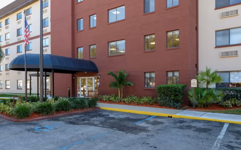 Entrance and facade of a multi-story senior living building with a covered awning, flagpoles, landscaping, and parking spaces.
