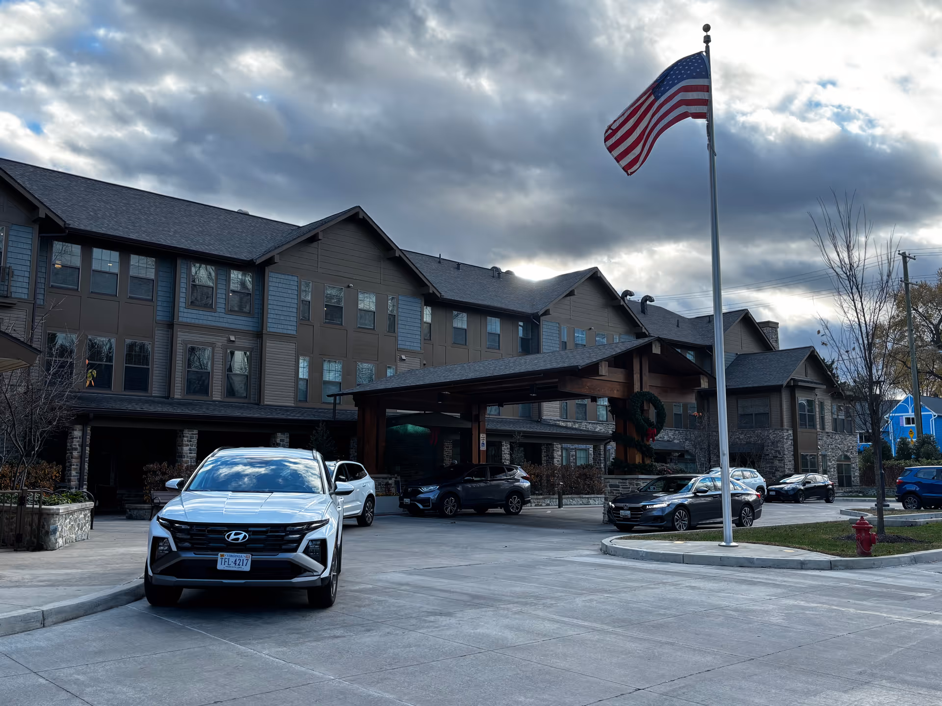 Exterior view of a senior living facility building with a covered entrance. Several cars are parked in front of the building. An American flag is flying on a flagpole near the entrance. The sky is partly cloudy.