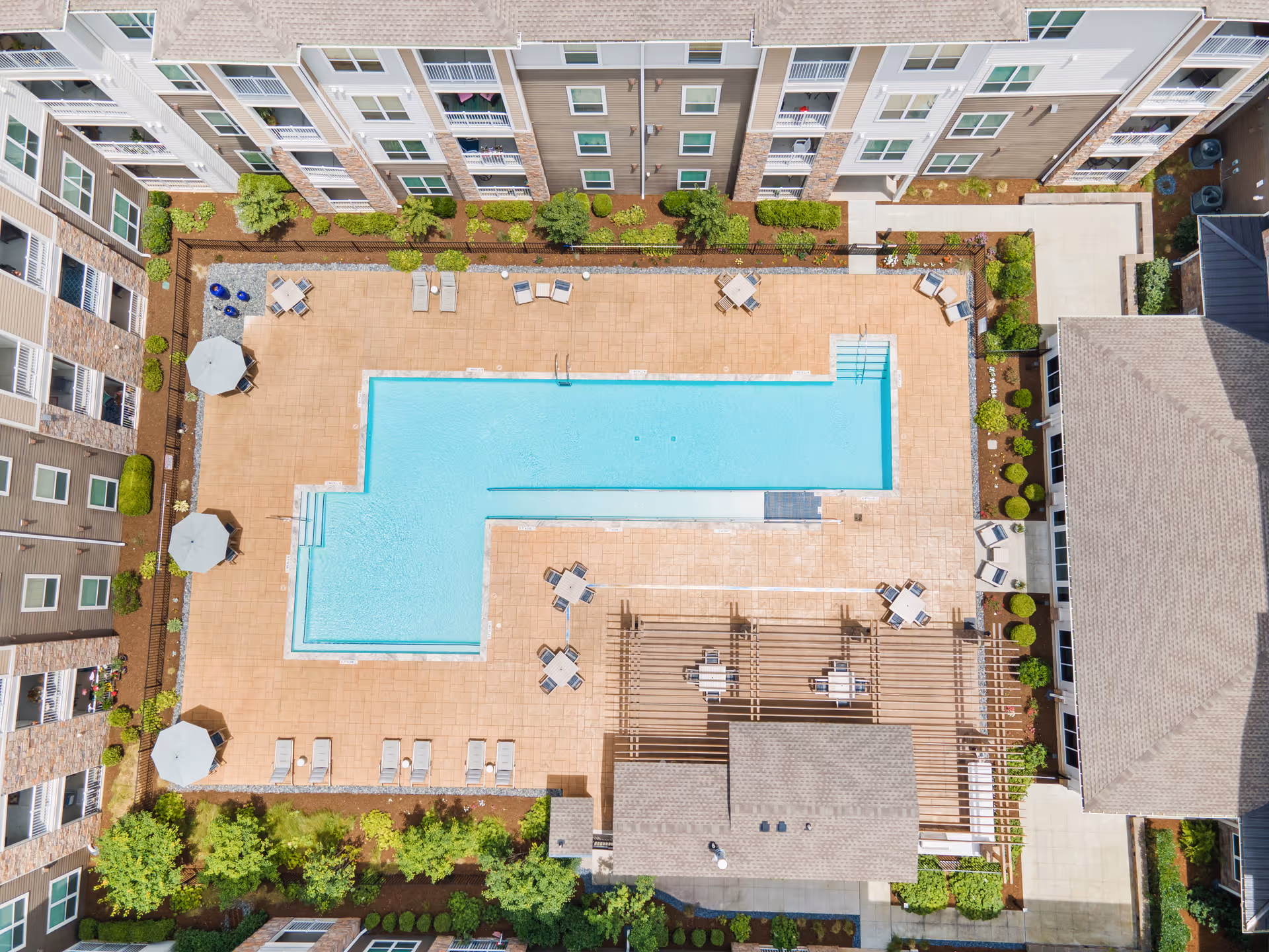 Aerial view of a rectangular outdoor swimming pool surrounded by a spacious patio area with lounge chairs, tables with umbrellas, and greenery. The pool area is enclosed by a multi-story residential building with balconies overlooking the pool.