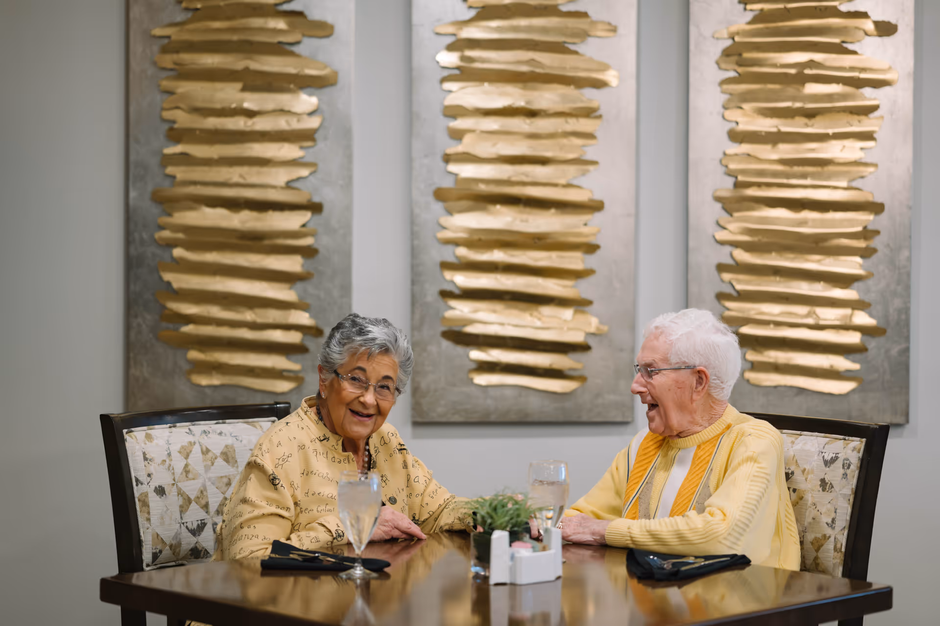 Two older adults smiling and talking at a dining table with glasses and decorative gold wall art behind them.