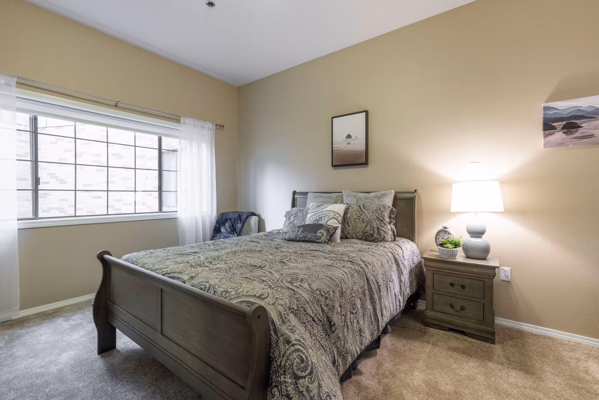 A cozy bedroom with a large window covered by sheer white curtains. The room features a wooden bed with a patterned comforter and multiple pillows. Next to the bed is a wooden nightstand with a lamp, a small plant, and a clock. Two framed pictures hang on the beige walls.