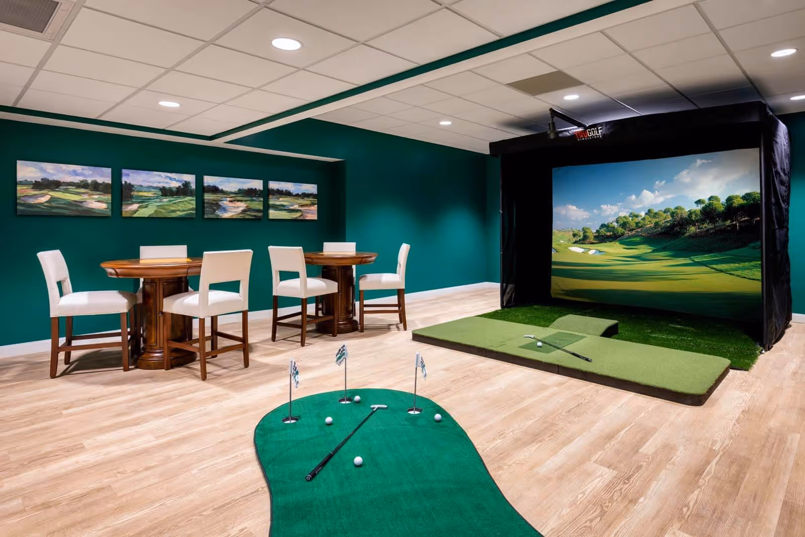 Indoor recreational room with a golf simulator setup, including a large screen displaying a golf course, a putting green mat with golf balls and a putter, and two wooden tables with white cushioned chairs against a teal wall adorned with four landscape paintings.