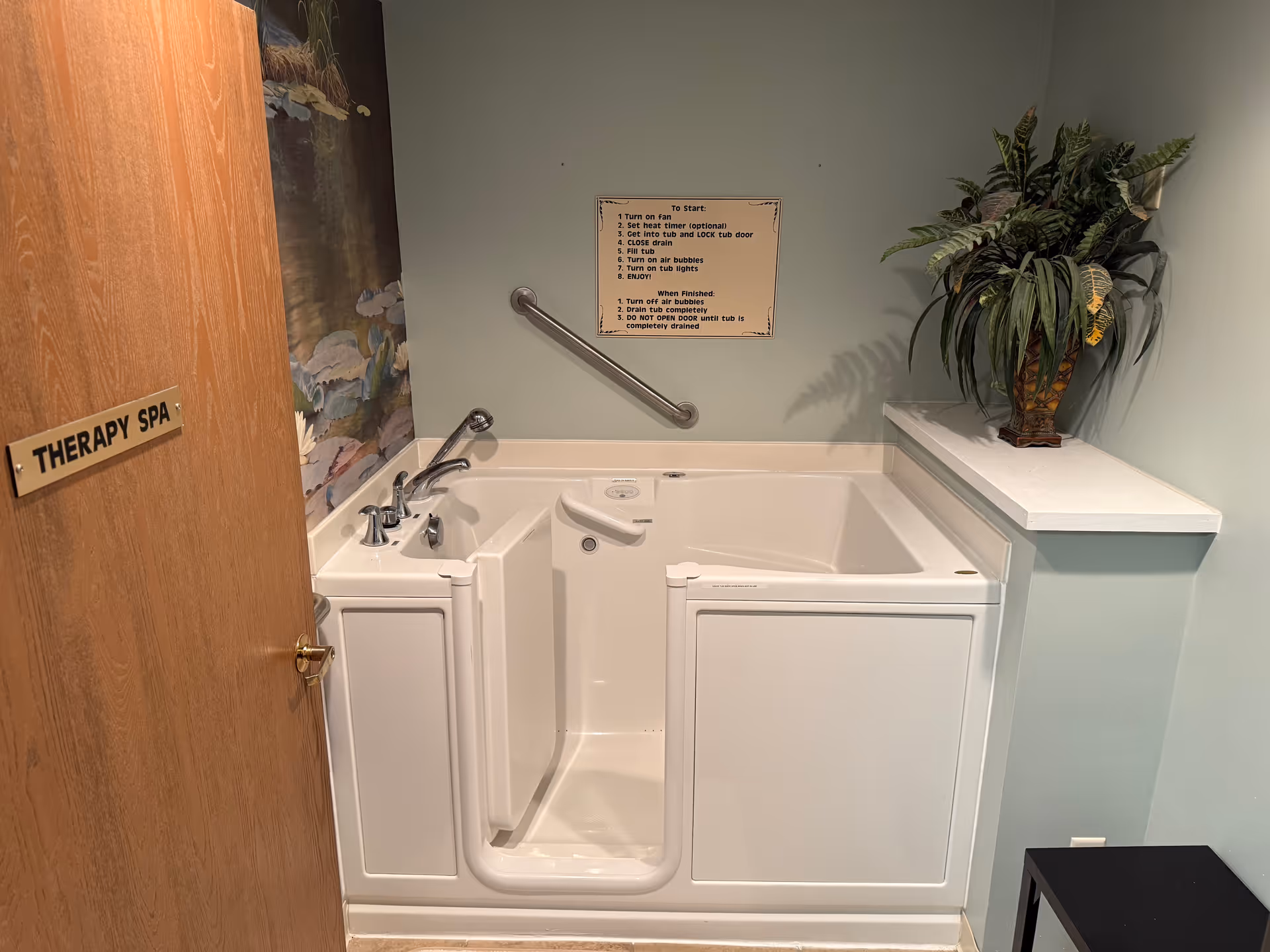 A therapy spa room featuring a white walk-in bathtub with handrails and faucet controls. There is a wooden door on the left with a sign that reads 'THERAPY SPA'. Above the bathtub, instructions for use are posted on the wall. A potted plant sits on a small ledge to the right of the tub, and a small black stool is positioned on the floor nearby.