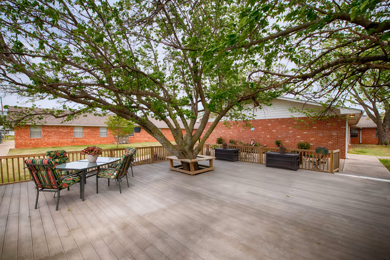 Spacious outdoor wooden deck with a large tree growing through the center, patio table and chairs, planters, and a brick building in the background.