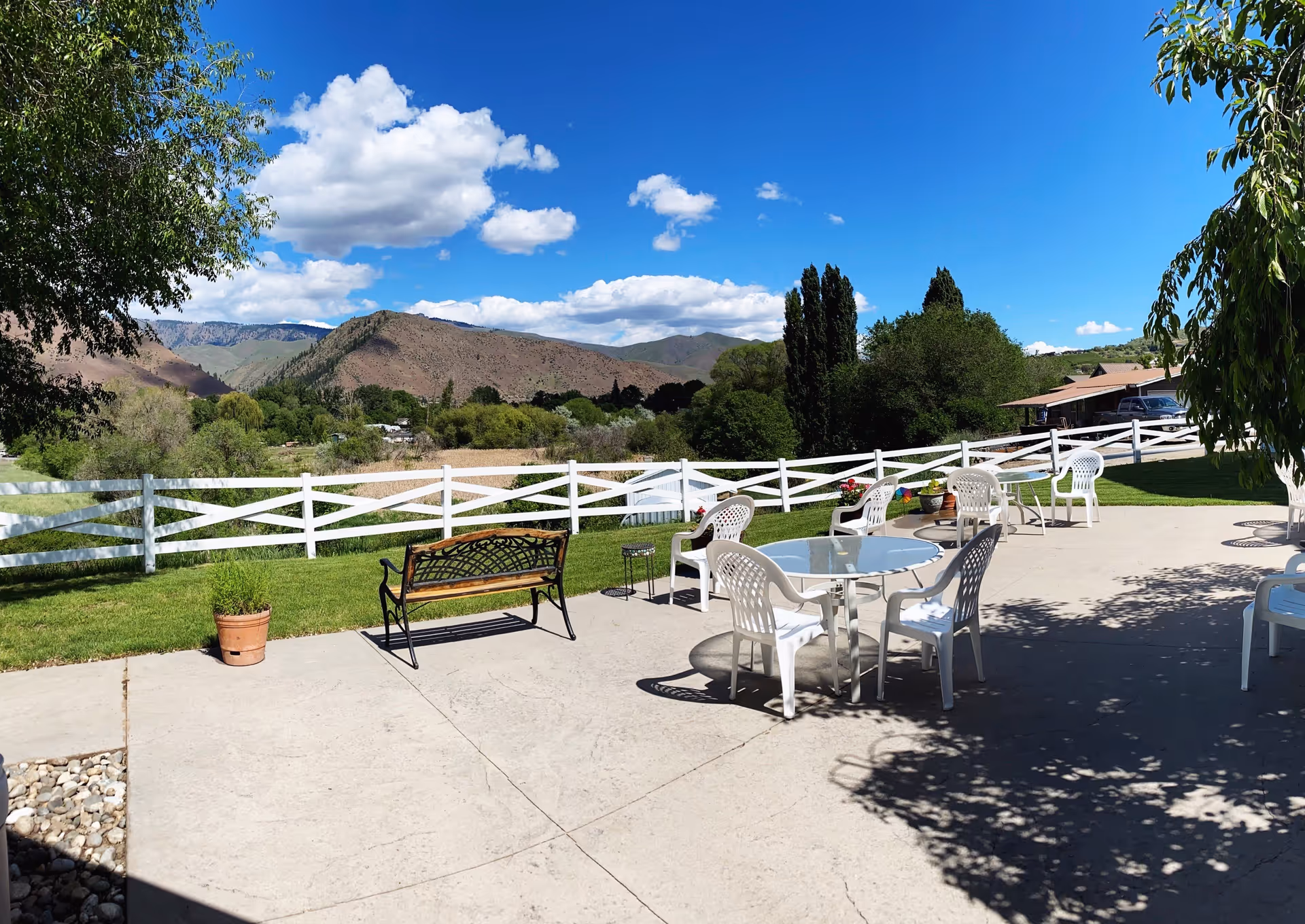 Patio with white plastic chairs and a glass table, a bench and potted plants overlooking a white fence, green lawn and distant mountains under a blue sky.