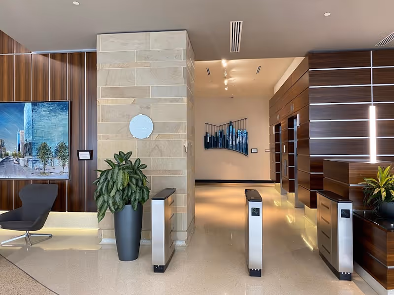 Modern interior lobby with wood panels, turnstiles, a large plant, and a wall-mounted screen.