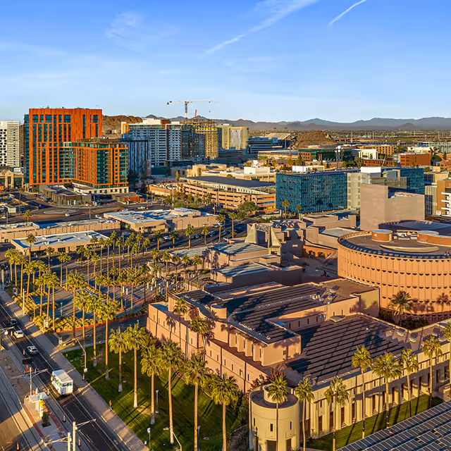 Aerial view of a cityscape featuring modern buildings, palm trees lining the streets, and distant mountains under a clear blue sky.