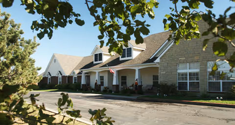 Exterior view of a senior living facility building with a covered porch, multiple windows, and a driveway. The building is surrounded by greenery and trees under a clear blue sky.