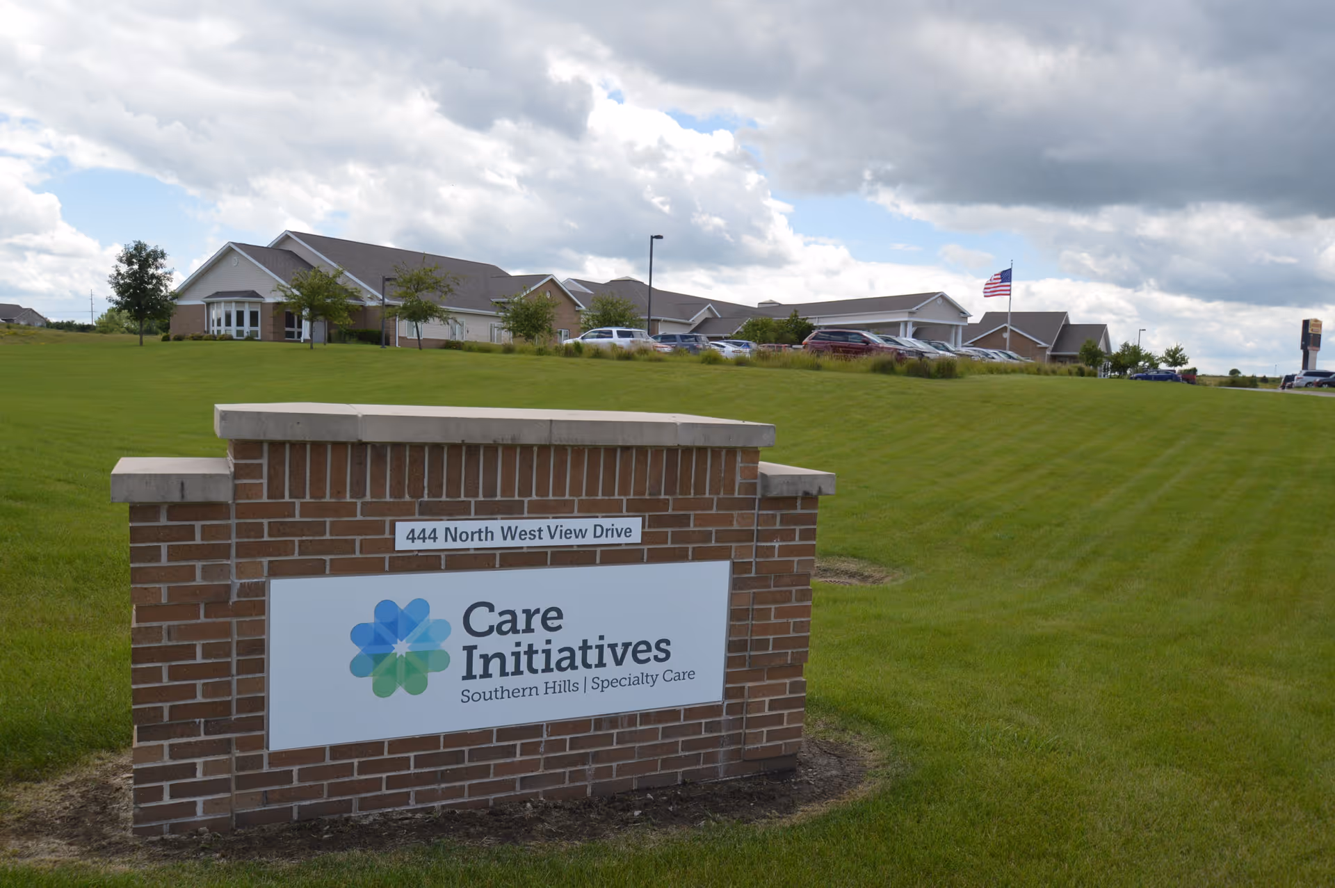 Exterior view of a senior living facility named Care Initiatives Southern Hills Specialty Care, showing a brick sign with the address 444 North West View Drive in the foreground and the building with parked cars and an American flag in the background under a partly cloudy sky.