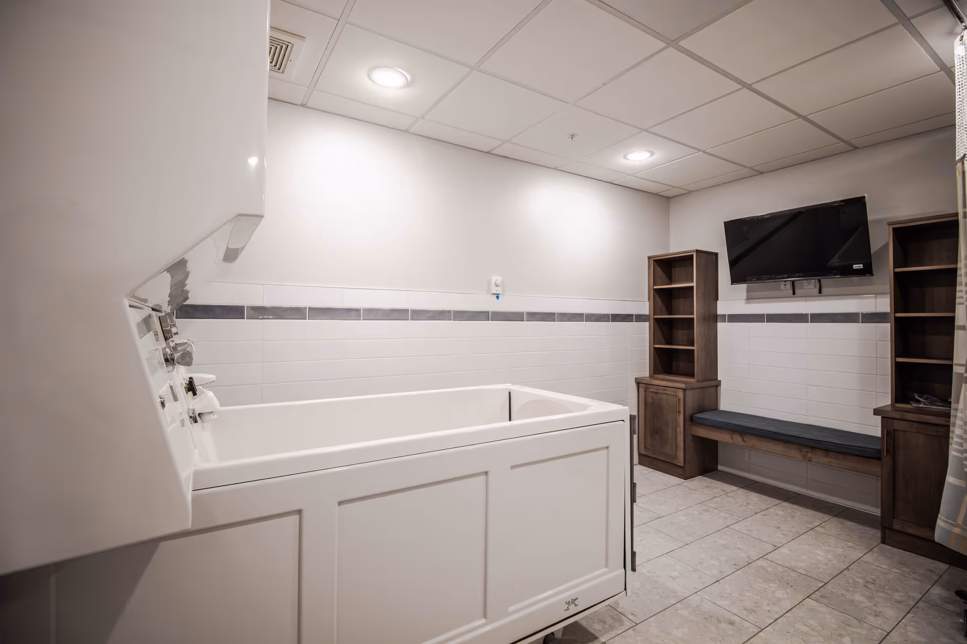 Bright accessible bathroom with a large white tub, tiled walls, bench seating, and cabinets.