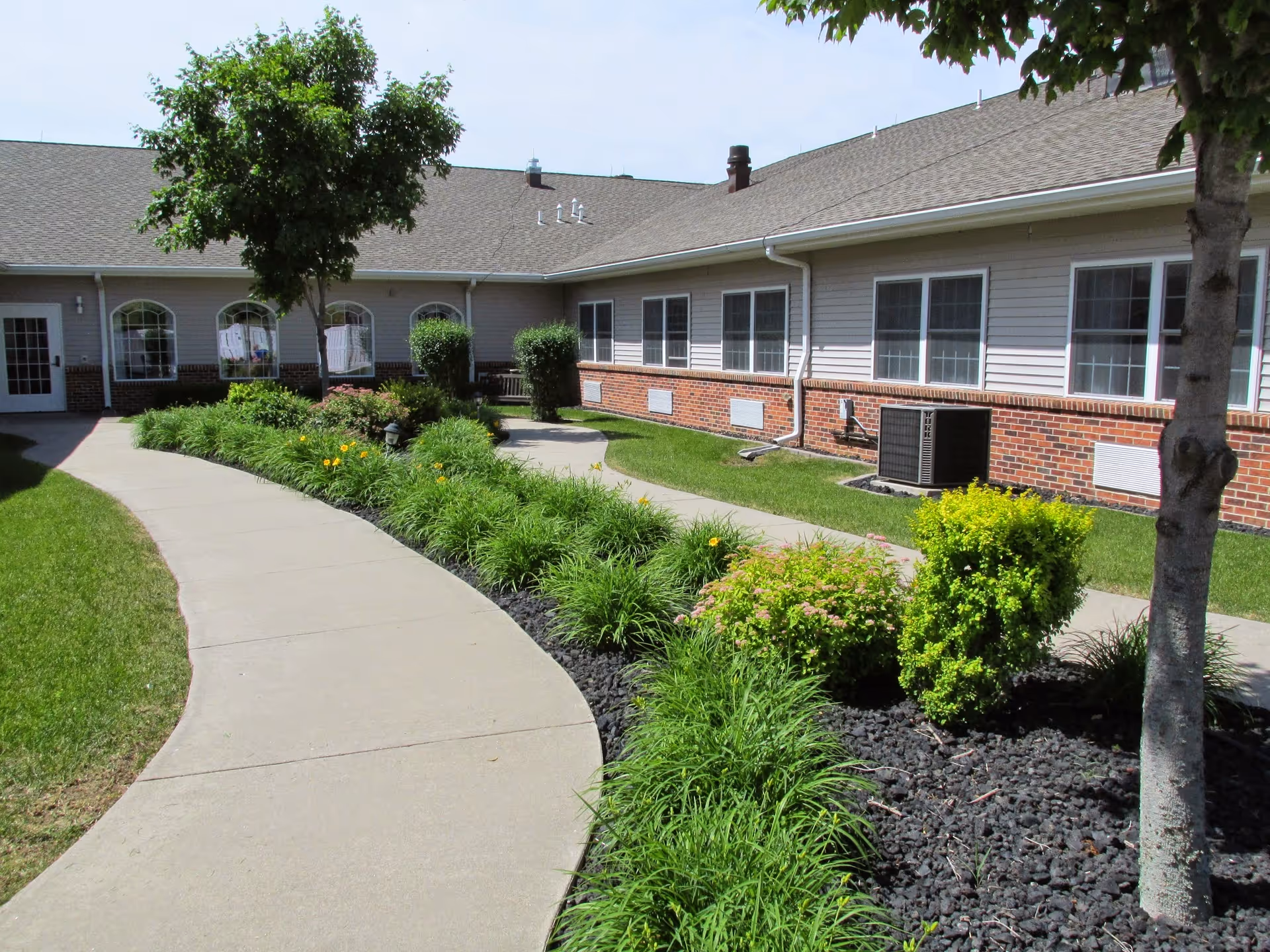 Curved concrete walkway winds through landscaped lawns and flowerbeds in front of a single-story brick-and-siding senior living building.