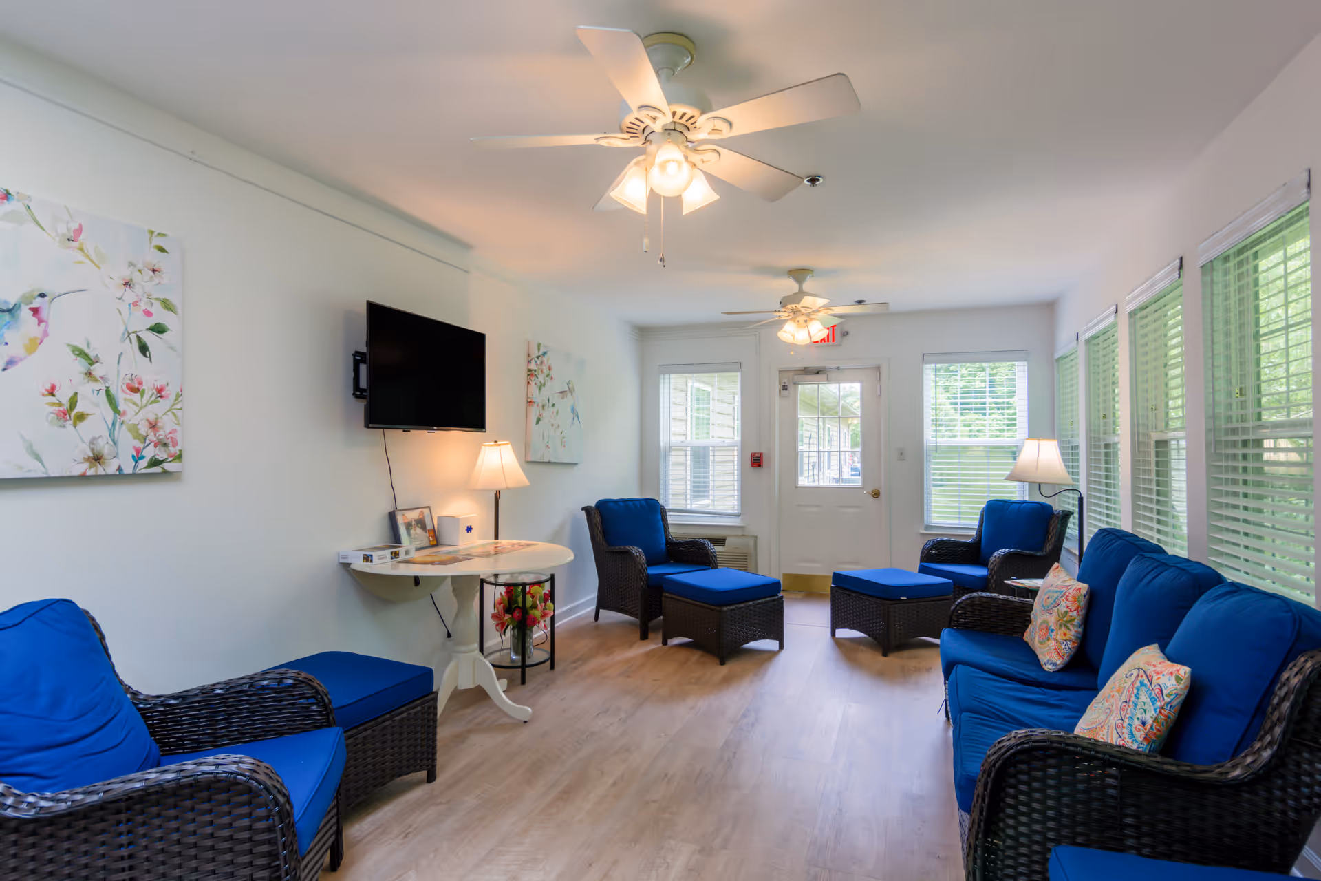 Bright common living room with blue cushioned wicker seating, tables, and ceiling fans.