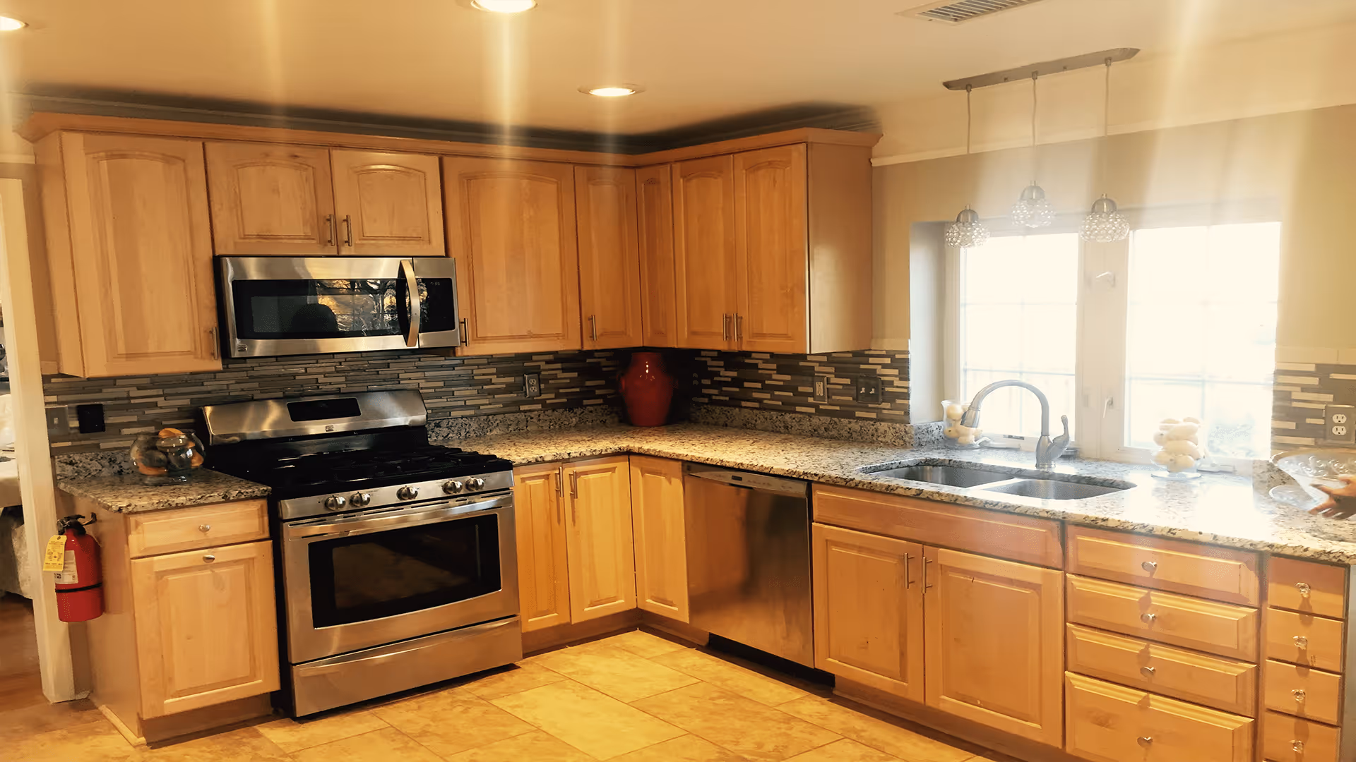 A bright kitchen with wooden cabinets, granite countertops, a stainless steel oven and microwave, a dishwasher, and a double sink under a window with three pendant lights hanging above.