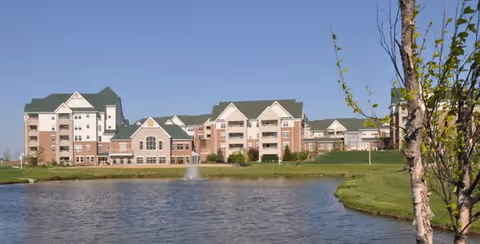 Multi-story senior living building with green roofs viewed across a pond with a small fountain and grassy landscaping.