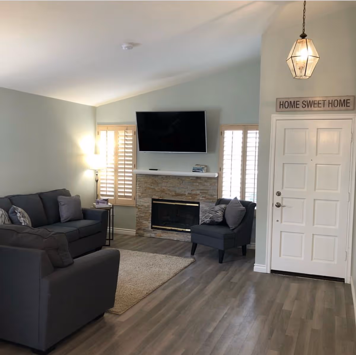Cozy living room with gray sofas, a stone fireplace topped by a wall-mounted TV, shuttered windows, and a white front door with a "Home Sweet Home" sign.