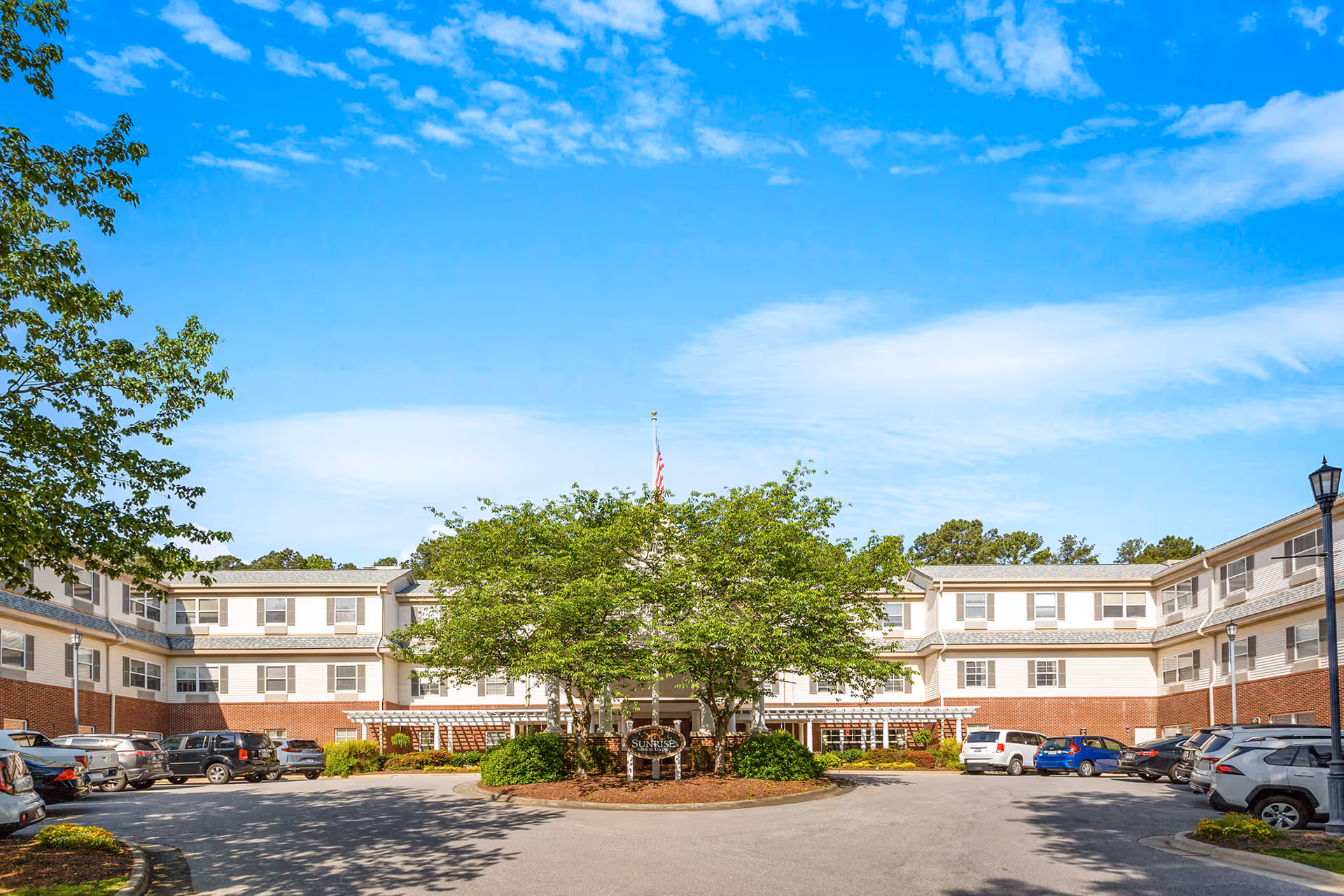 Sunrise at North Hills exterior view of a U-shaped senior living facility with trees and cars.