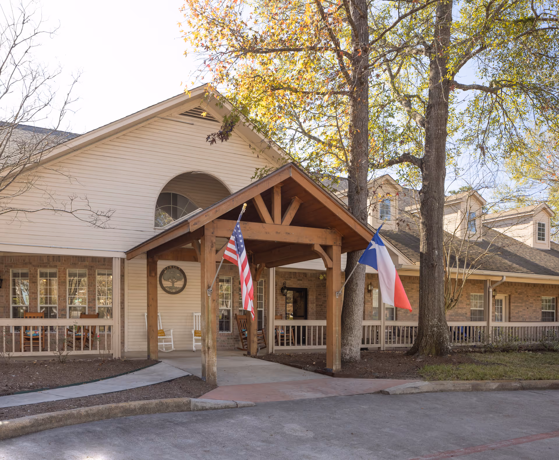 Front entrance of a senior living building with a wooden covered portico, American and Texas flags, rocking chairs on the porch, and large trees.