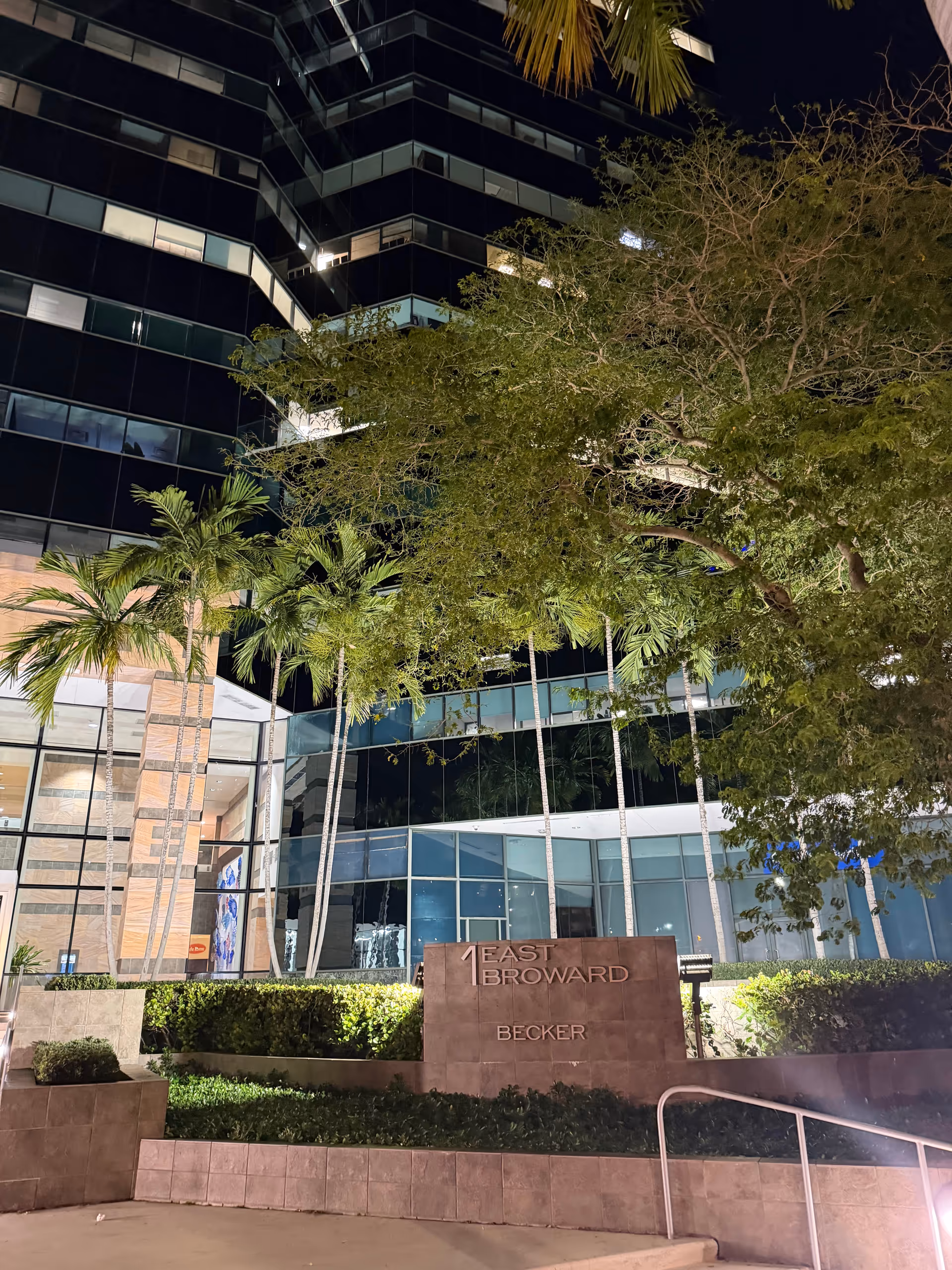 Night exterior of a modern glass building with palm trees and an East Broward sign