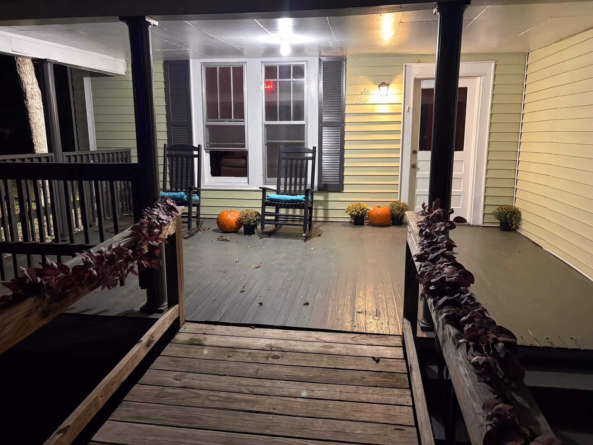 Covered front porch with rocking chairs, pumpkins, and potted plants in front of a light-green house.