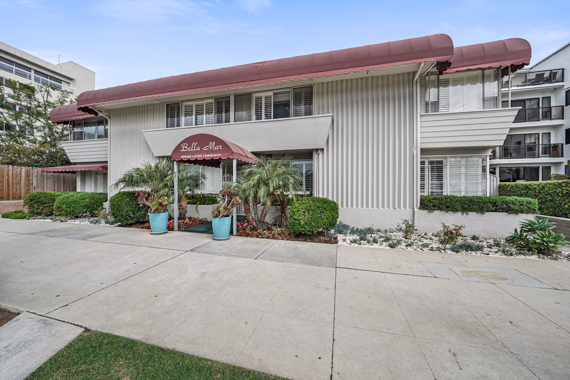 Exterior view of a senior living community building named Bella Mar with beige walls and maroon awnings. The entrance is covered by a maroon awning with the text 'Bella Mar Senior Living Community'. There are potted plants and landscaping with bushes and flowers around the entrance. The sidewalk and street are visible in the foreground.