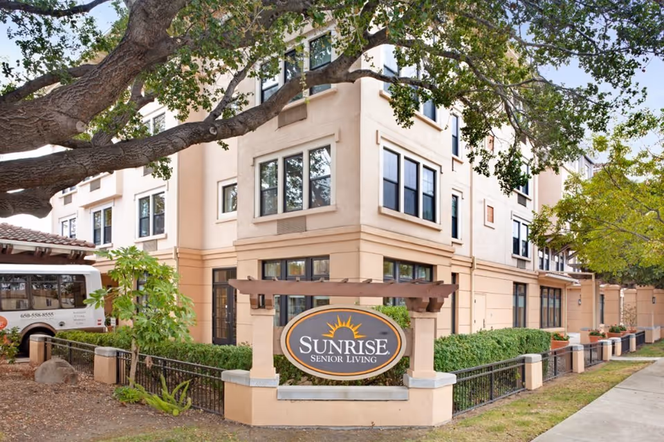 Exterior view of Sunrise Senior Living facility in San Mateo, showing a multi-story beige building with multiple windows, a large tree in the foreground, and a sign with the Sunrise Senior Living logo near the entrance. A white shuttle van is parked on the left side.