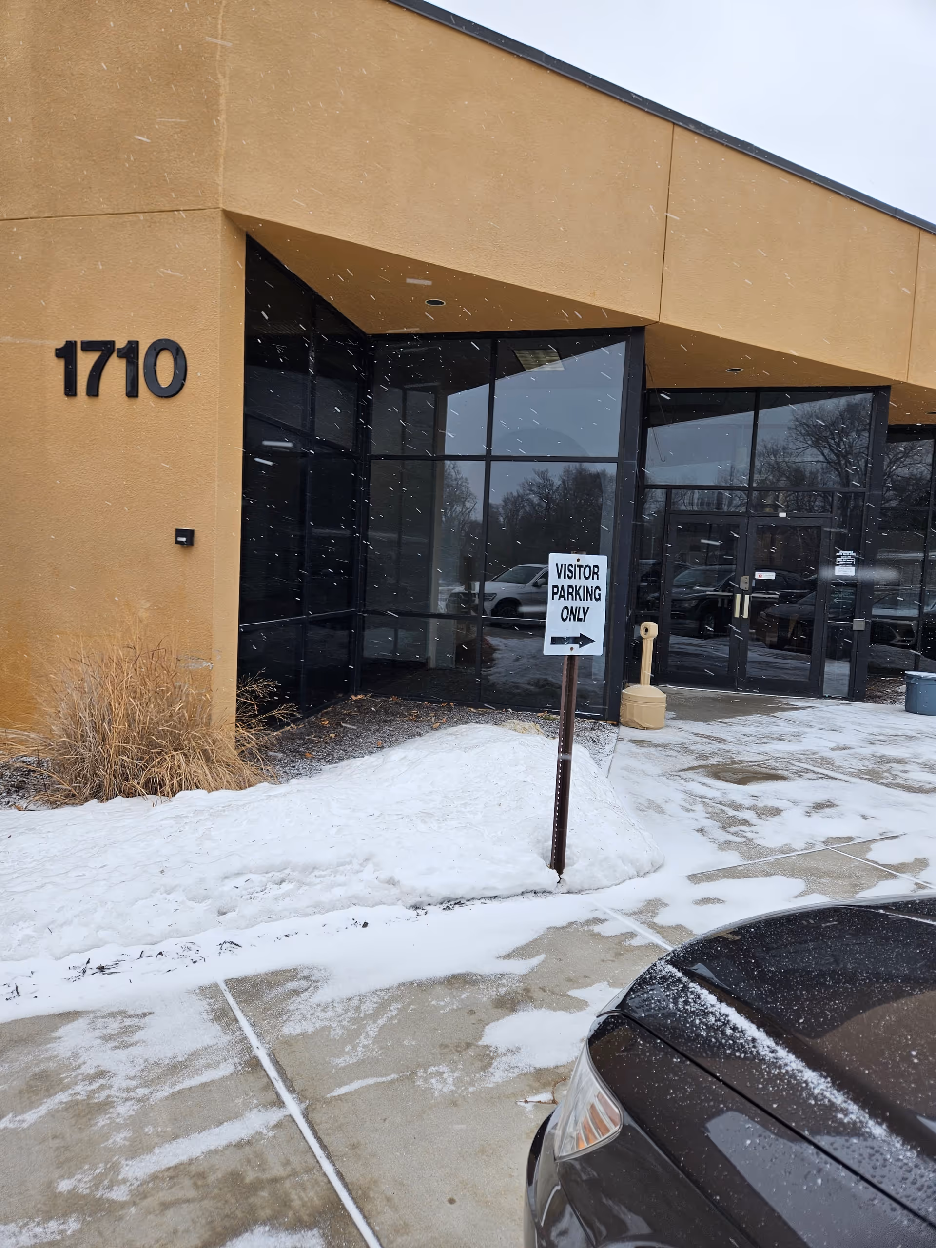 Exterior view of beige building entrance with glass doors and visitor parking sign in snow.