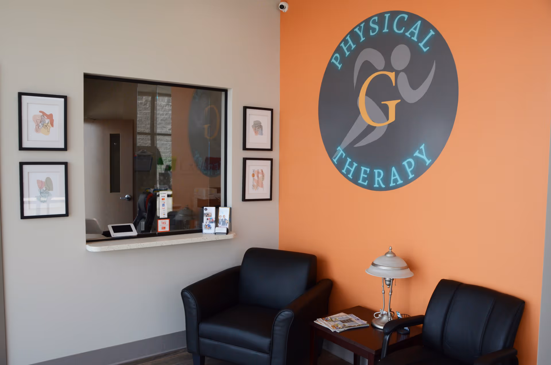 A modern therapy center seating area with orange logo wall, two black chairs, lamp, and magazines.