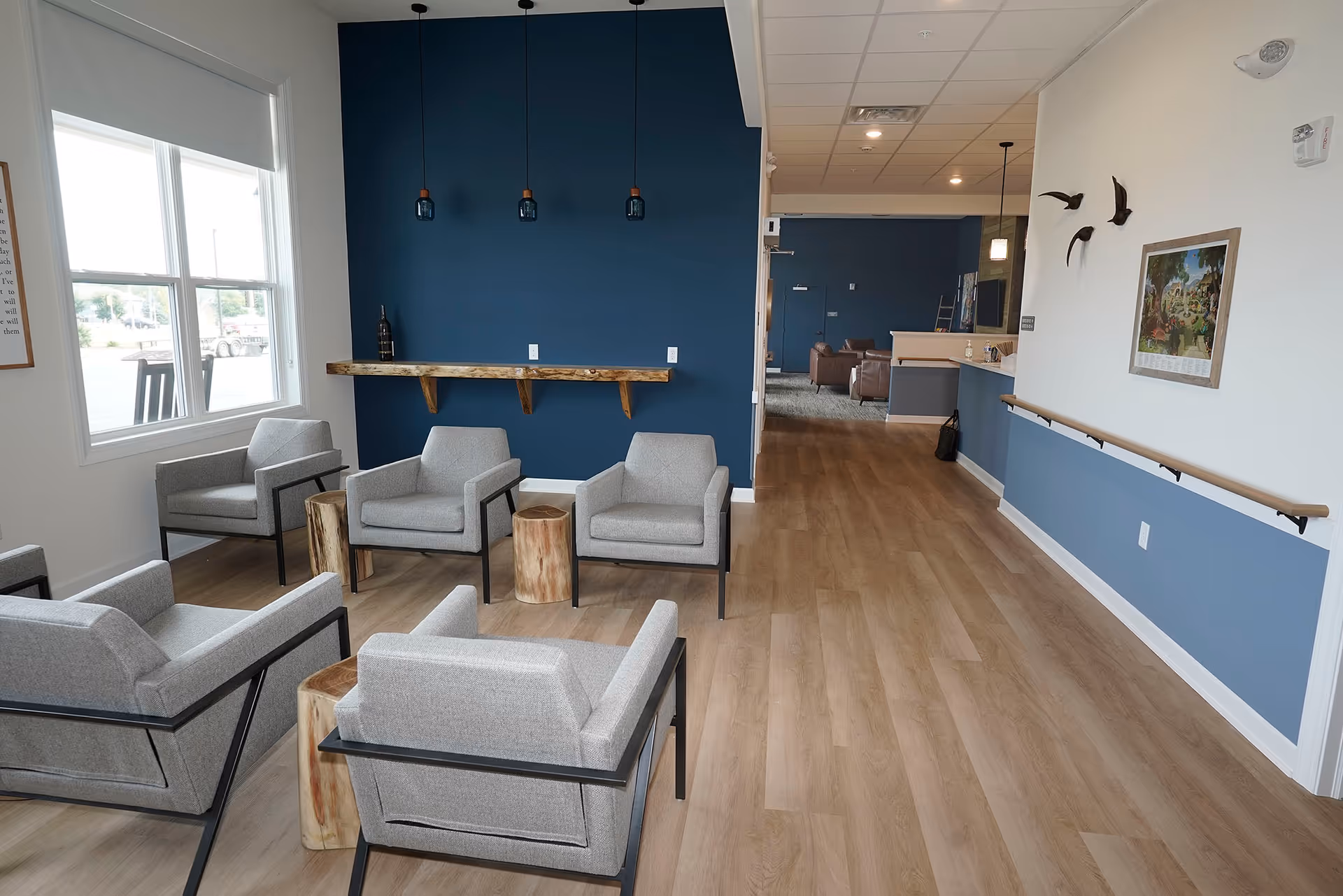 Lounge area with gray armchairs, wooden stools, blue accent wall, and a hallway in the background.