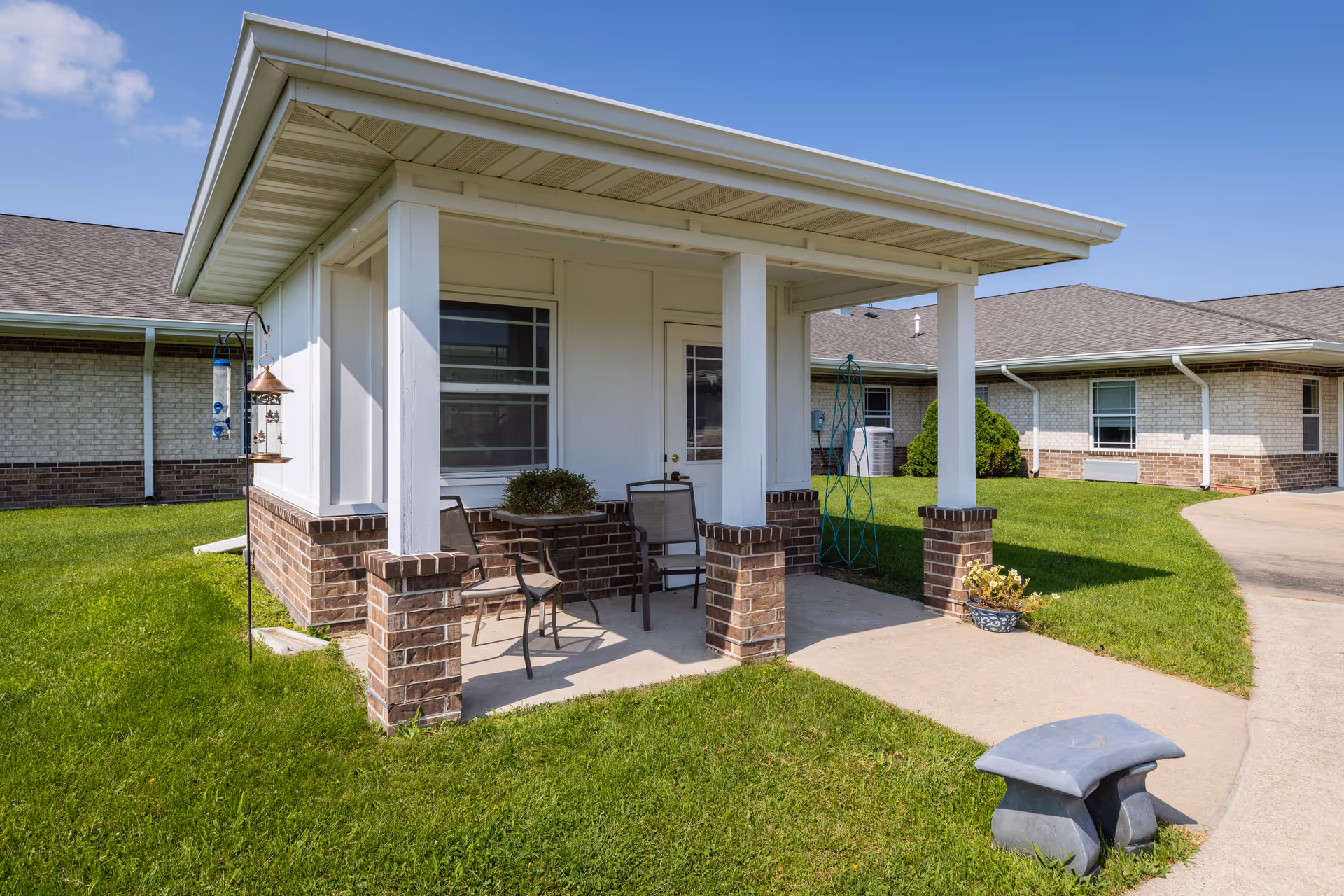 Covered brick porch with white pillars, chairs, and potted plants at the exterior of a single-story senior living building.