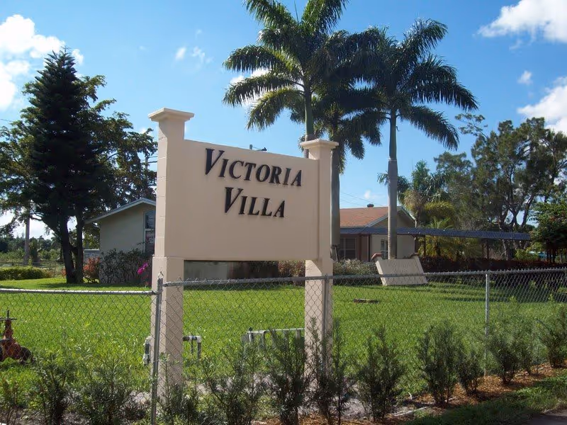 Front view of Victoria Villa sign with palm trees and a fenced lawn.