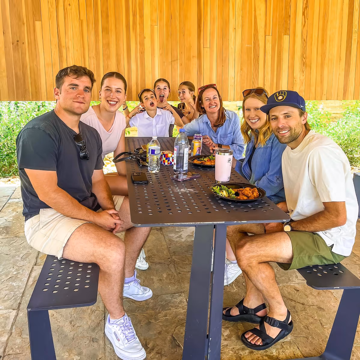 Group of adults sits around a metal outdoor table enjoying a meal under a wooden shelter.
