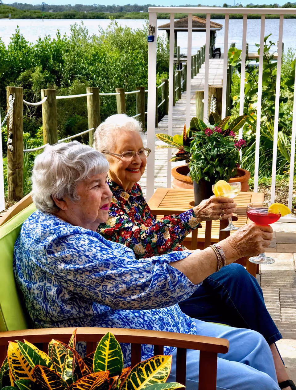 Two elderly women sitting outdoors on a patio near a waterfront, raising glasses with colorful drinks and lemon garnishes, surrounded by green plants and a wooden dock extending into the water in the background.