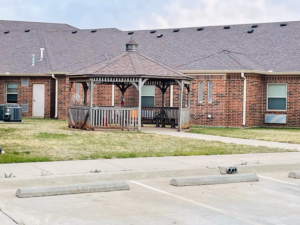Wooden gazebo on a grassy courtyard in front of a single-story brick senior living building with parking spaces in the foreground.