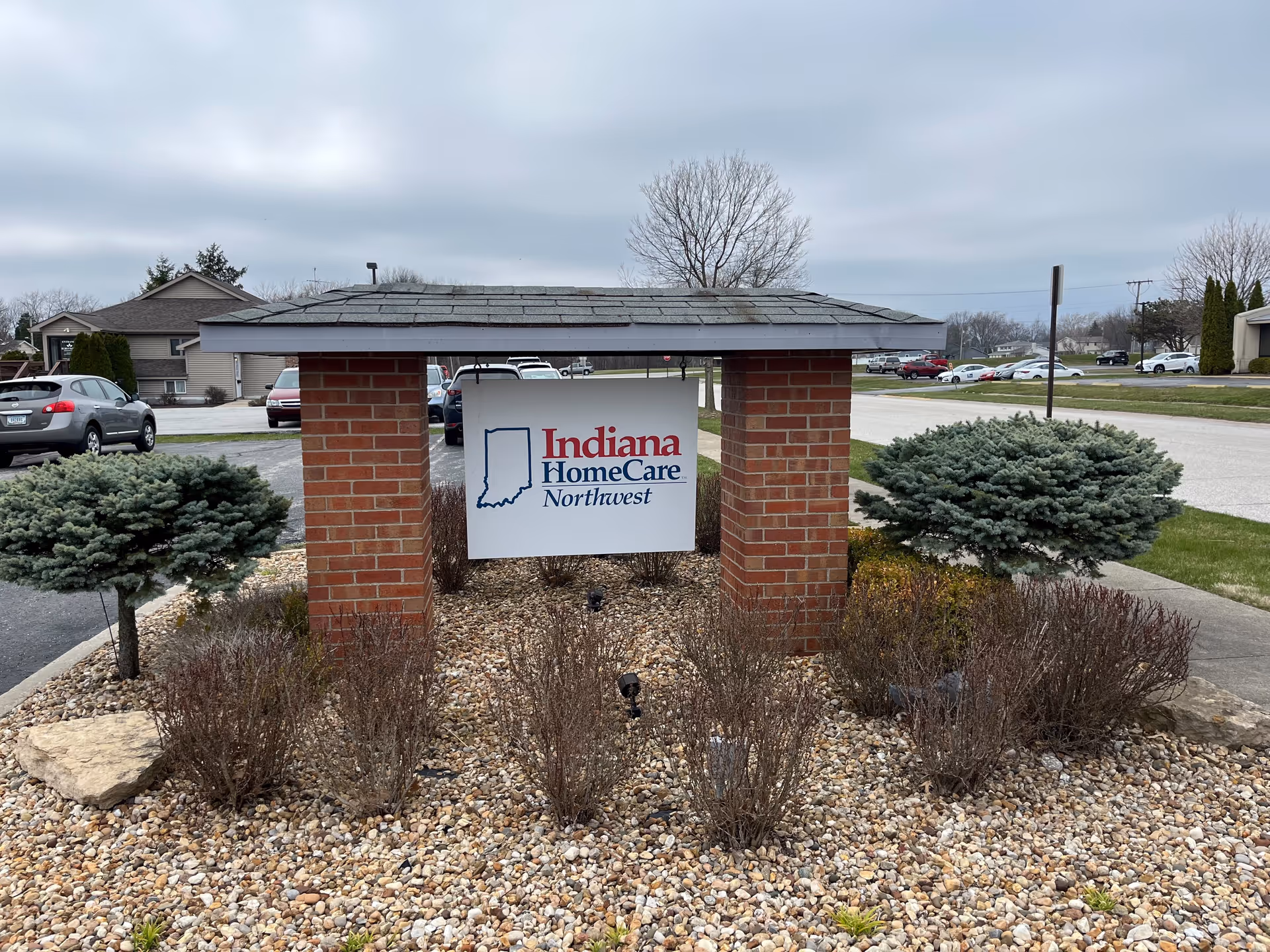 Brick sign shelter with Indiana HomeCare Northwest sign amid shrubs and a parking lot.