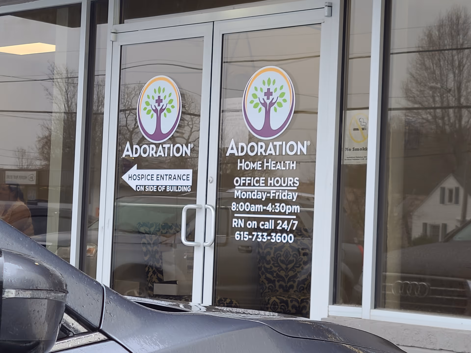 Glass entry doors display Adoration Hospice signs with a parked car outside.