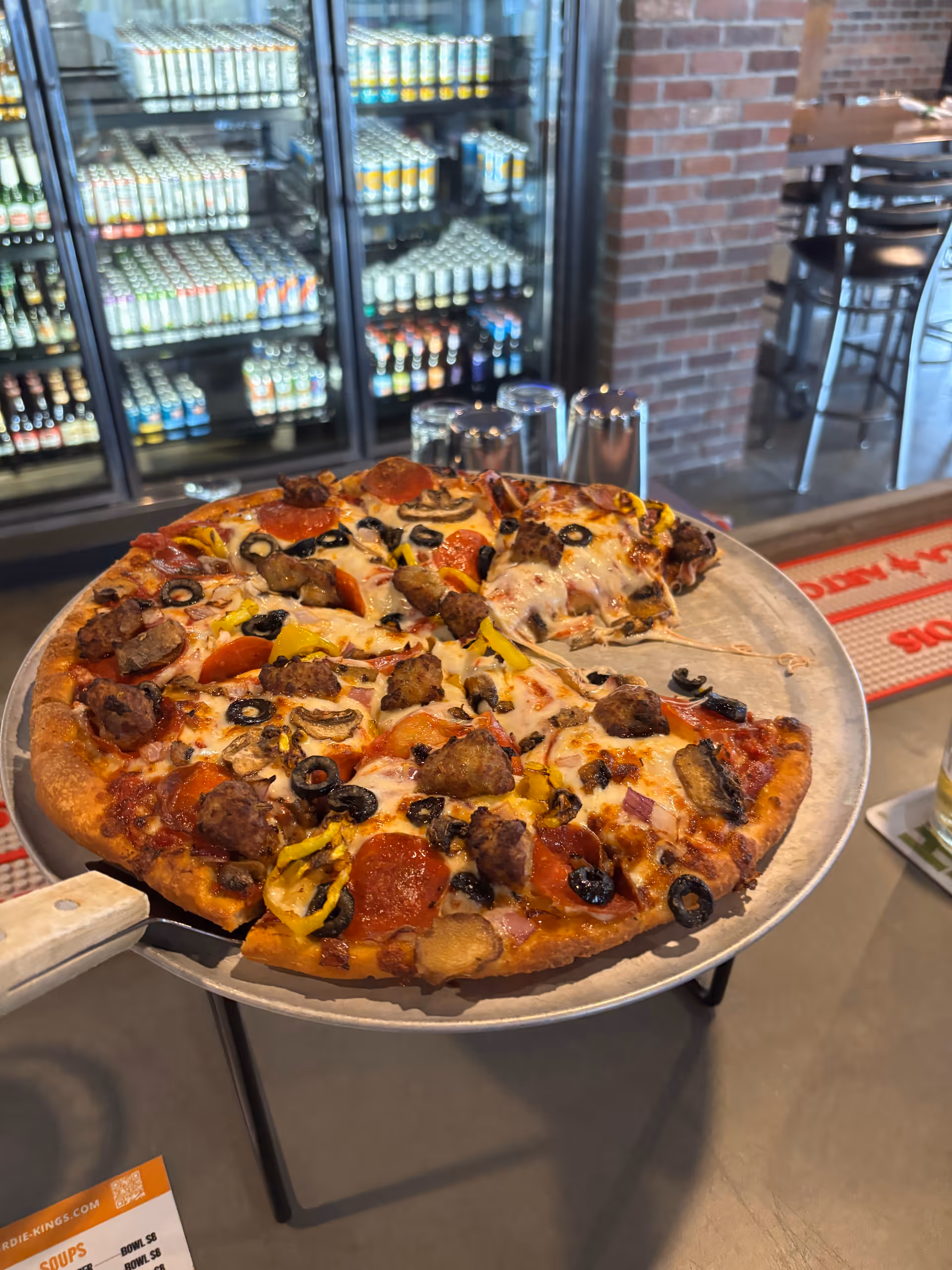 Large pepperoni pizza on a metal tray in a dining area with refrigerators in the background.