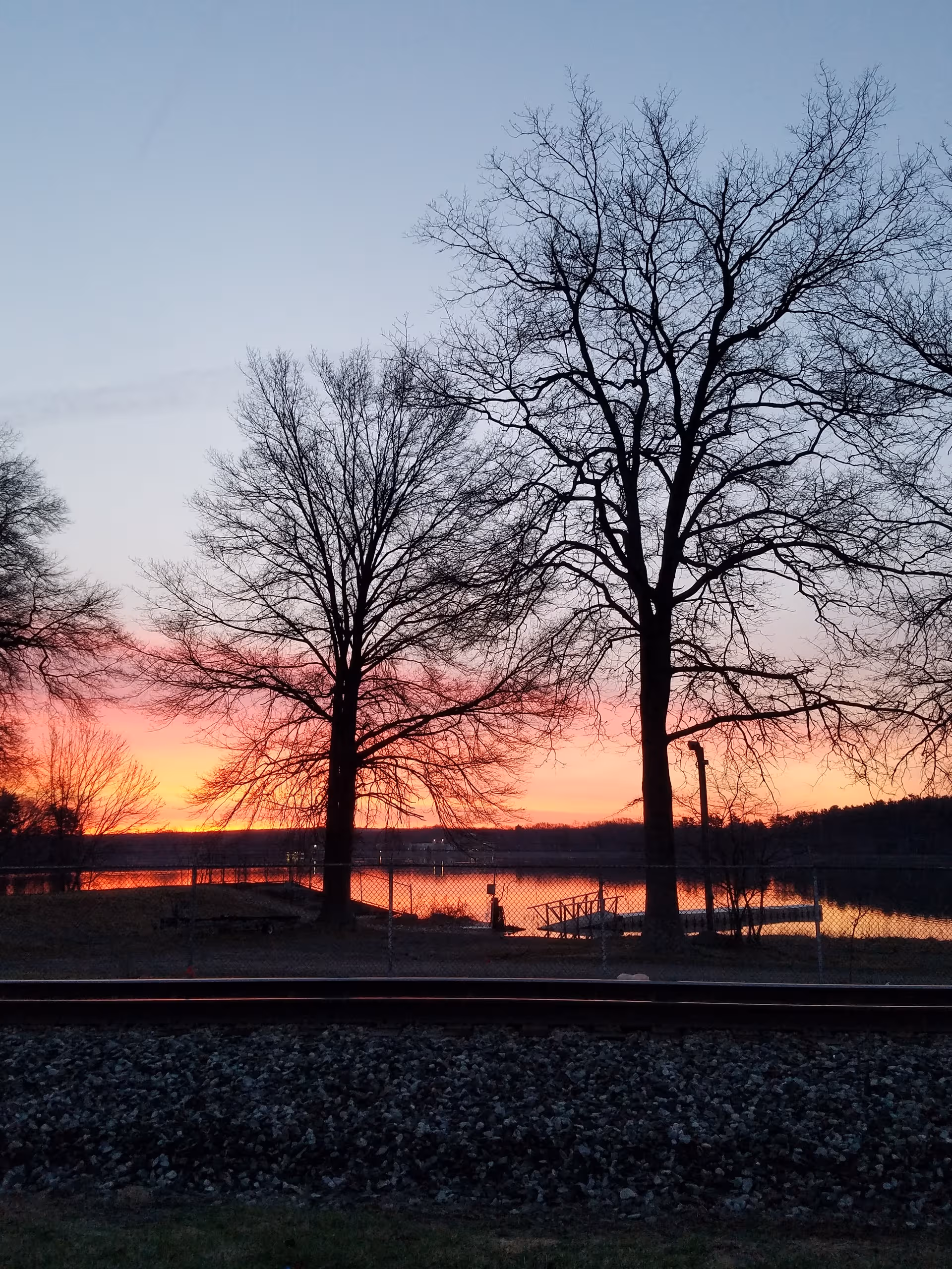 Sunset over a lake with bare trees and a rail line in the foreground.
